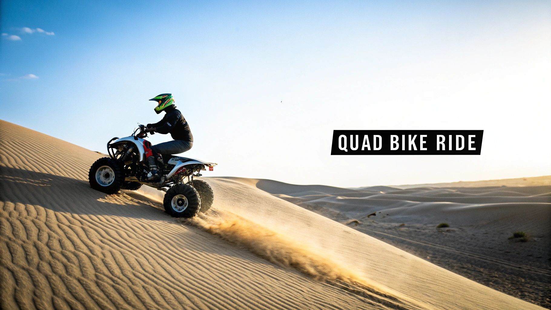 A person in a helmet rides a quad bike up a golden sand dune, kicking up dust in a vast desert.