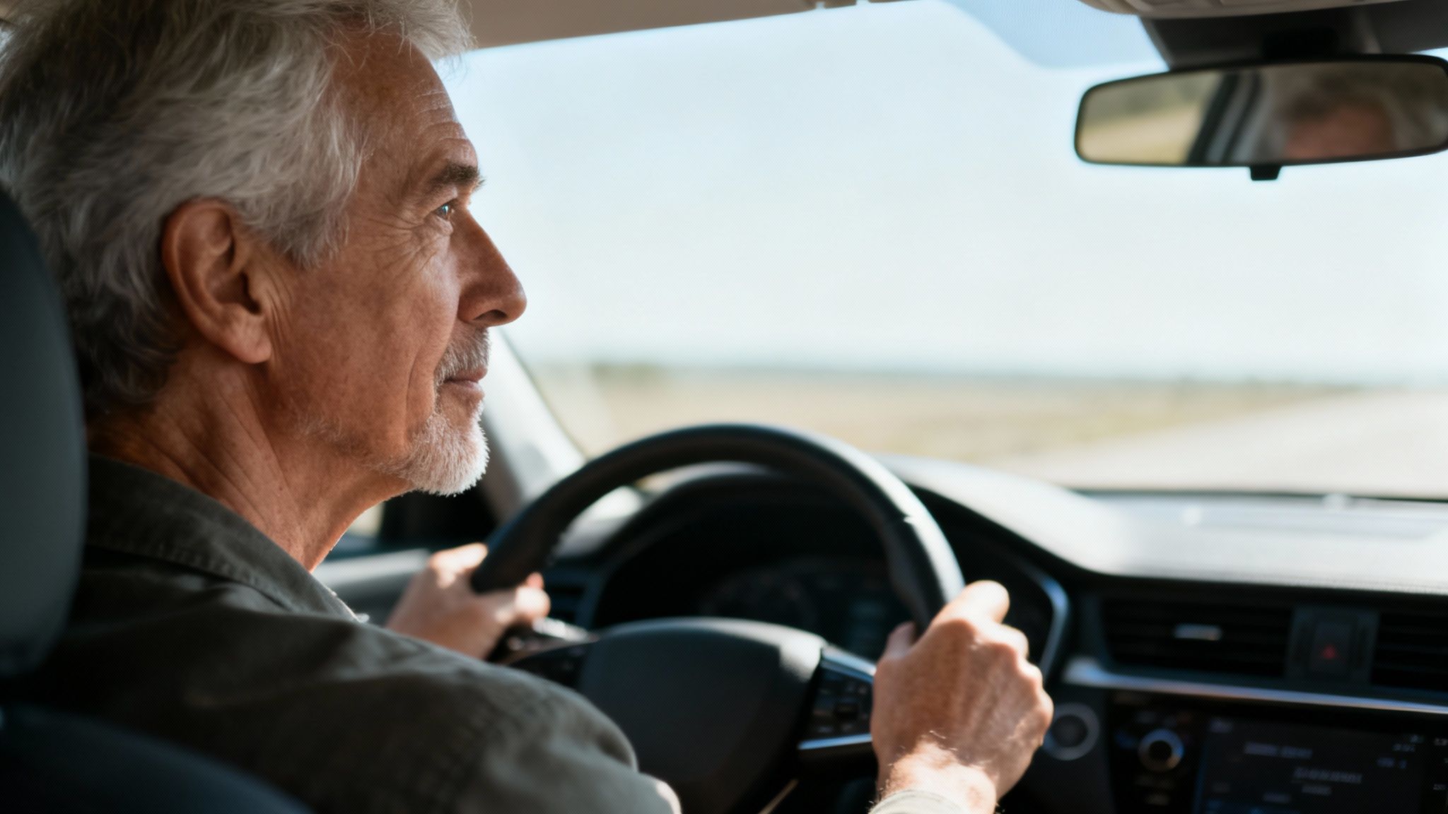 Vista de perfil de un hombre mayor con barba canosa, conduciendo un coche bajo el cielo azul.