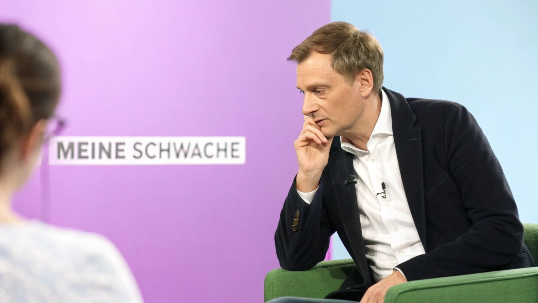 Nachdenklicher Mann im Interview, sitzt in einem grünen Sessel vor einer lila Wand mit der Aufschrift „MEINE SCHWACHE“.