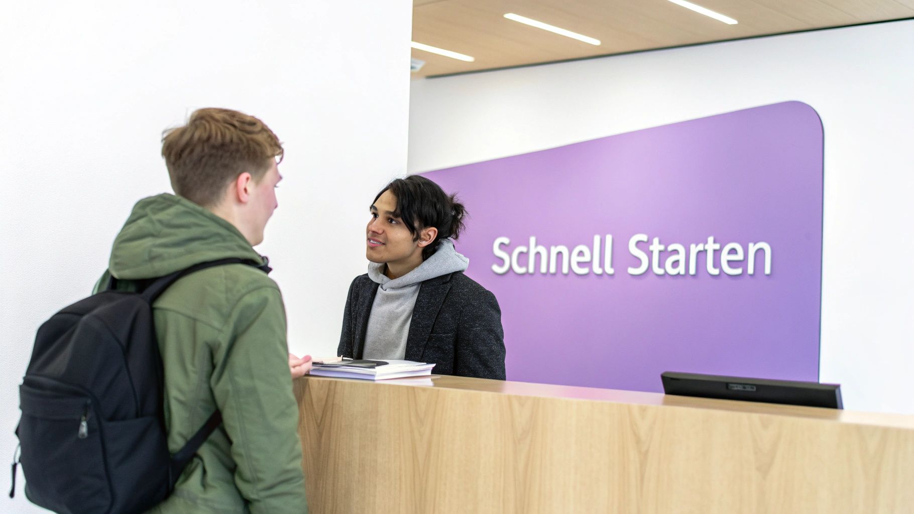 Zwei junge Männer an einem Empfangstresen, einer im Gespräch mit einem Mitarbeiter vor einer lila Wand mit der Aufschrift "Schnell Starten".