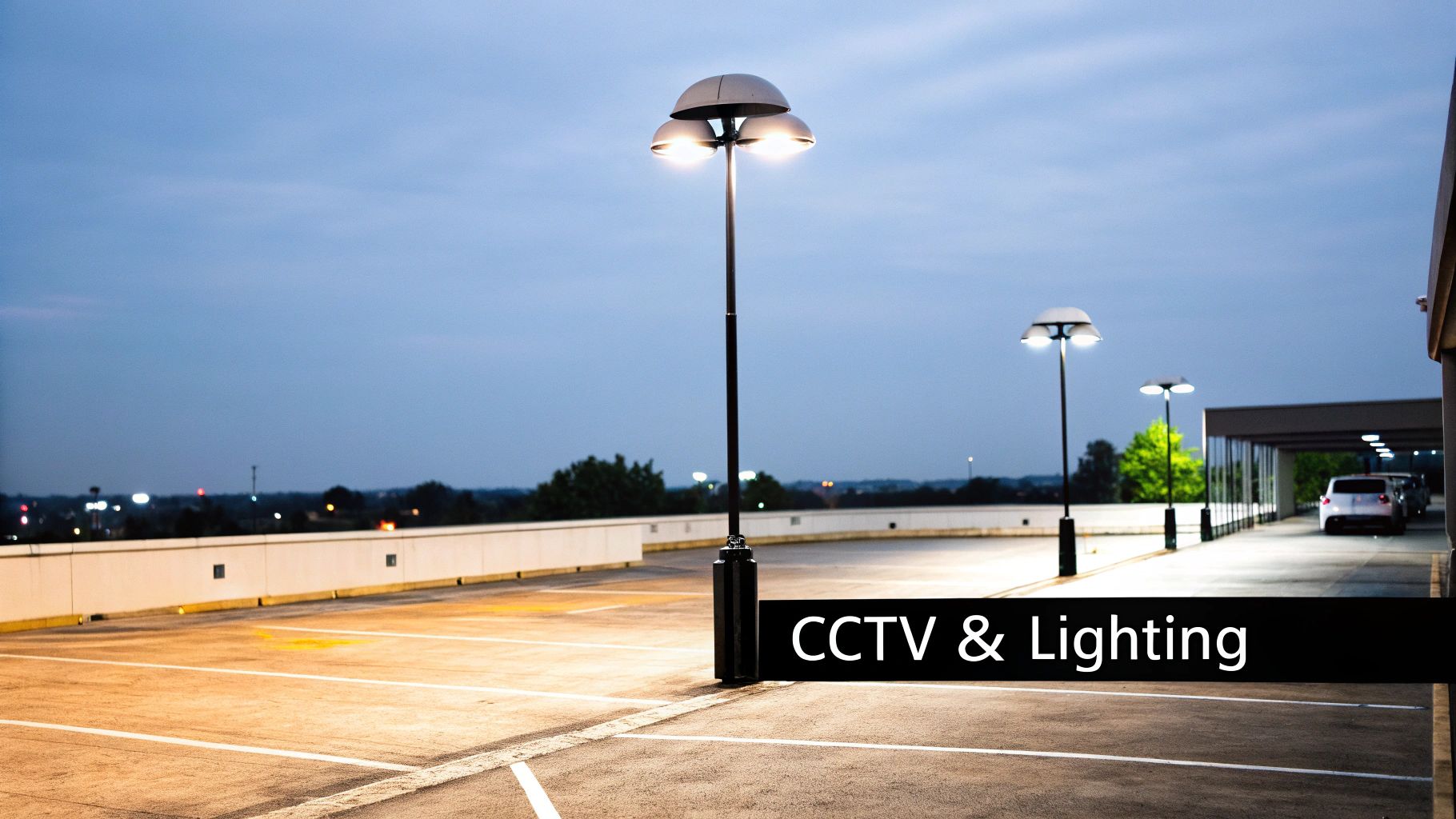 A modern CCTV camera mounted on a building, overlooking a well-lit car park at dusk.