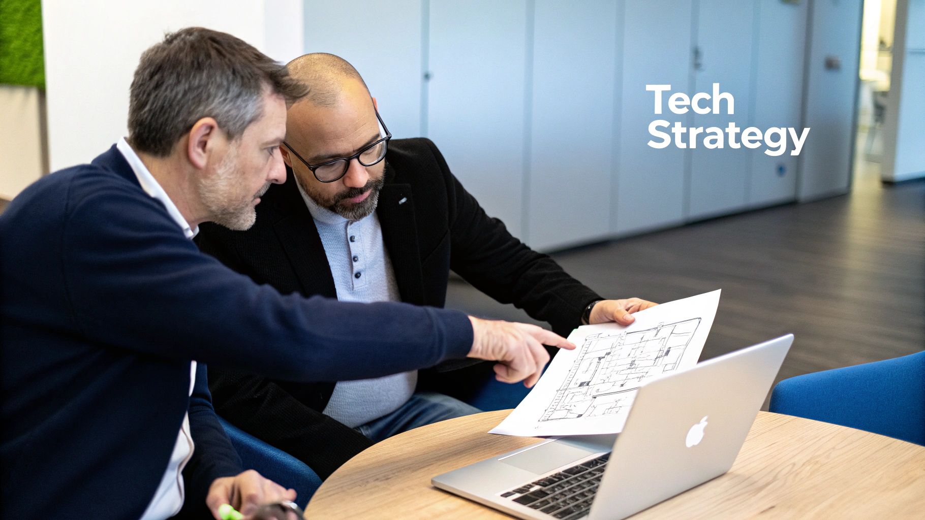 Two men discuss tech strategy, reviewing a blueprint with a laptop on a wooden table.