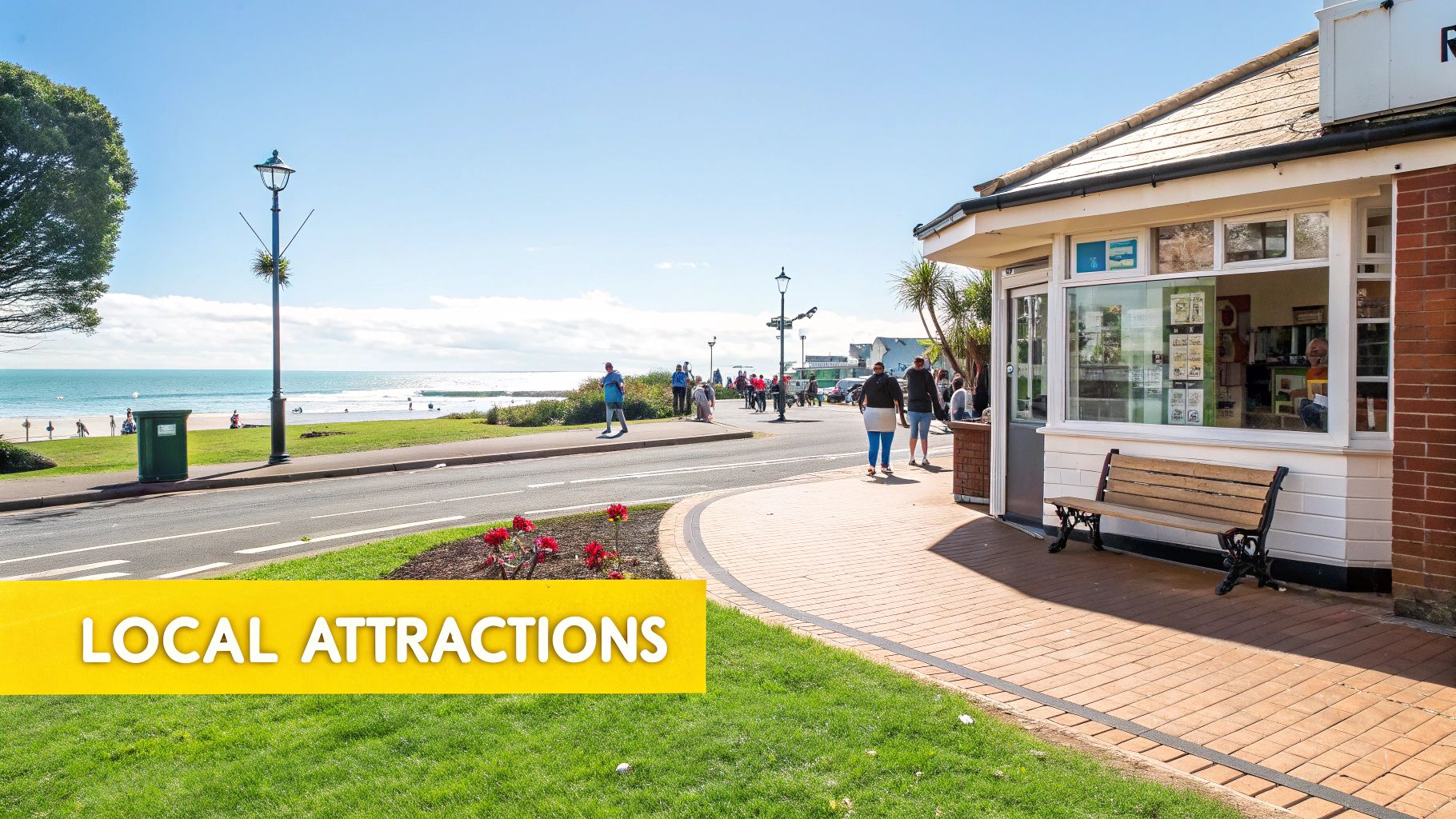 Sunny day at a coastal town with a beach, road, people walking, and a small building.