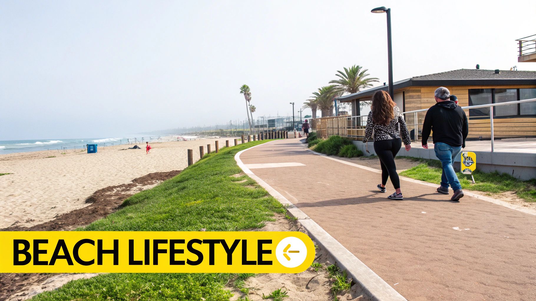 A sunny beach with ocean waves, a paved walking path, and people strolling by palm trees.