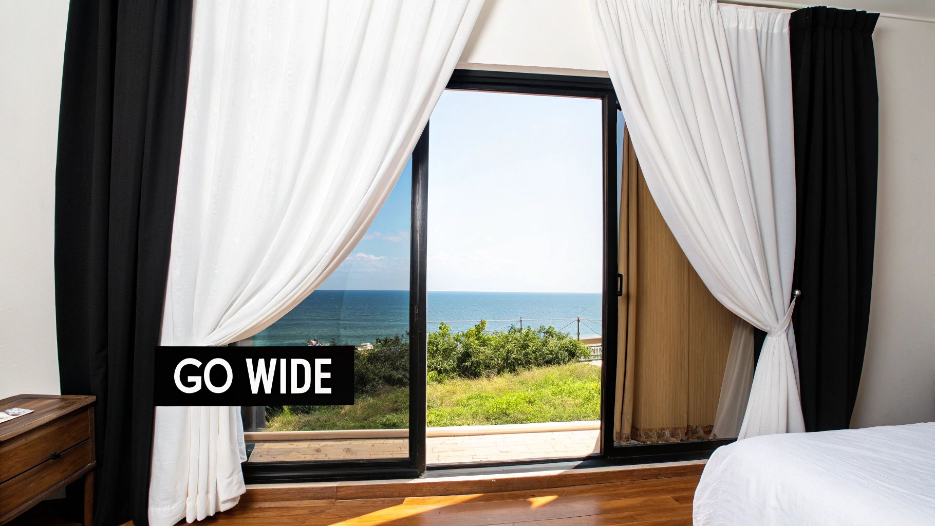 A hotel room with black and white drapes pulled back from a sliding glass door revealing an ocean view.