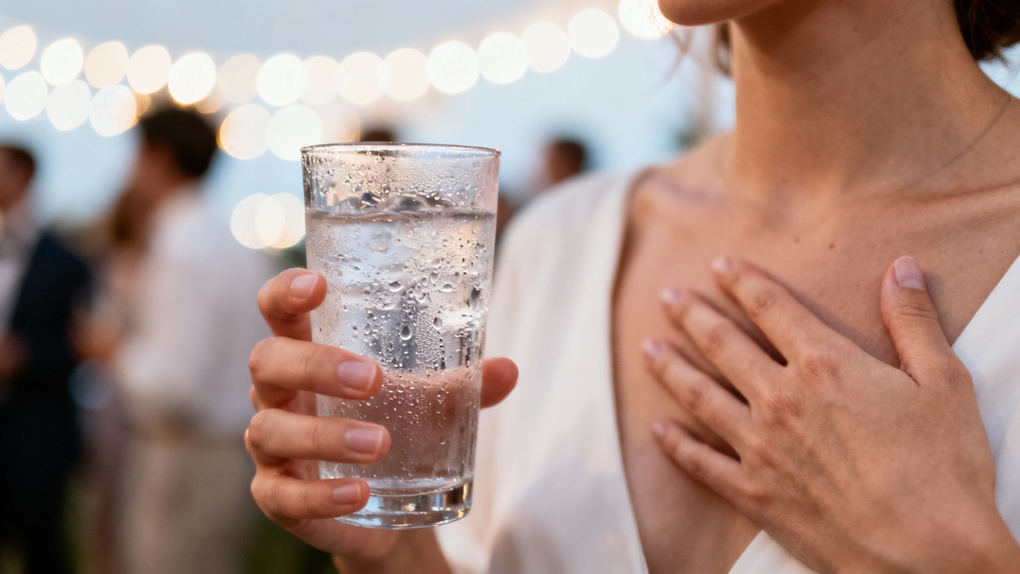 Close-up of a person holding a cold glass of water at a social gathering with blurred lights.