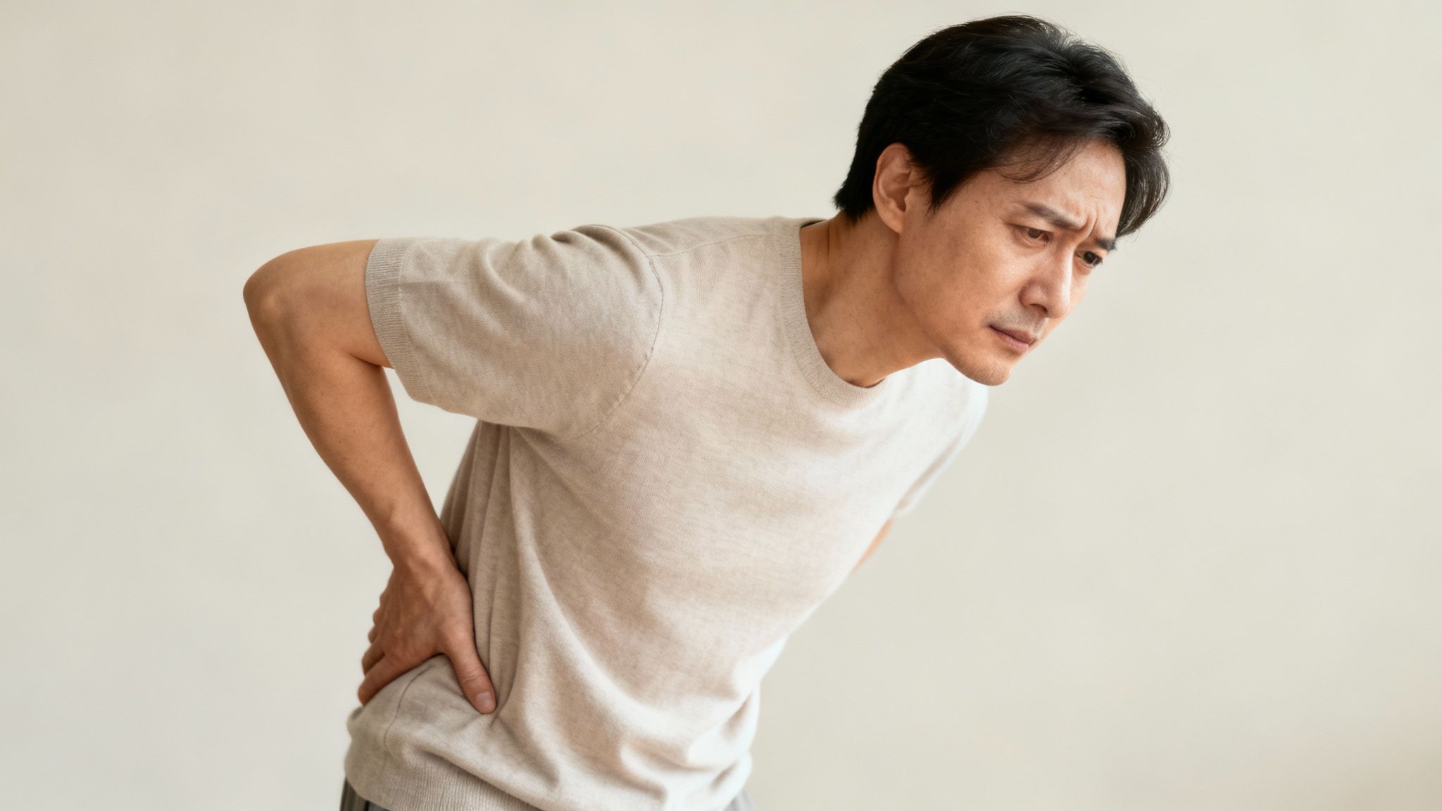 A distressed Asian man with dark hair holds his lower back, indicating pain, against a plain light background.