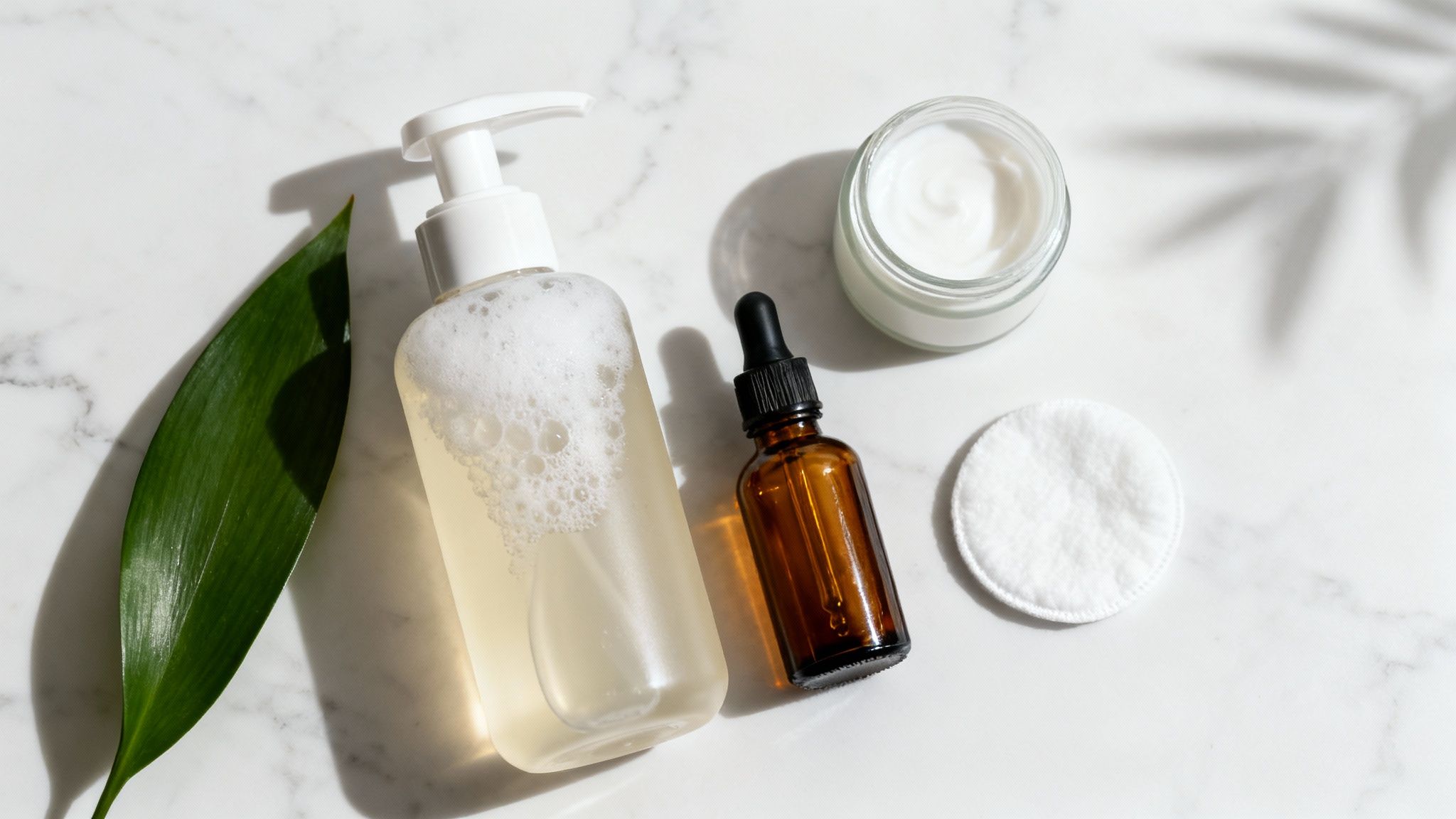 Prodotti per la cura della pelle sul marmo: detergente, siero, crema viso, dischetto di cotone e foglia verde alla luce del sole.