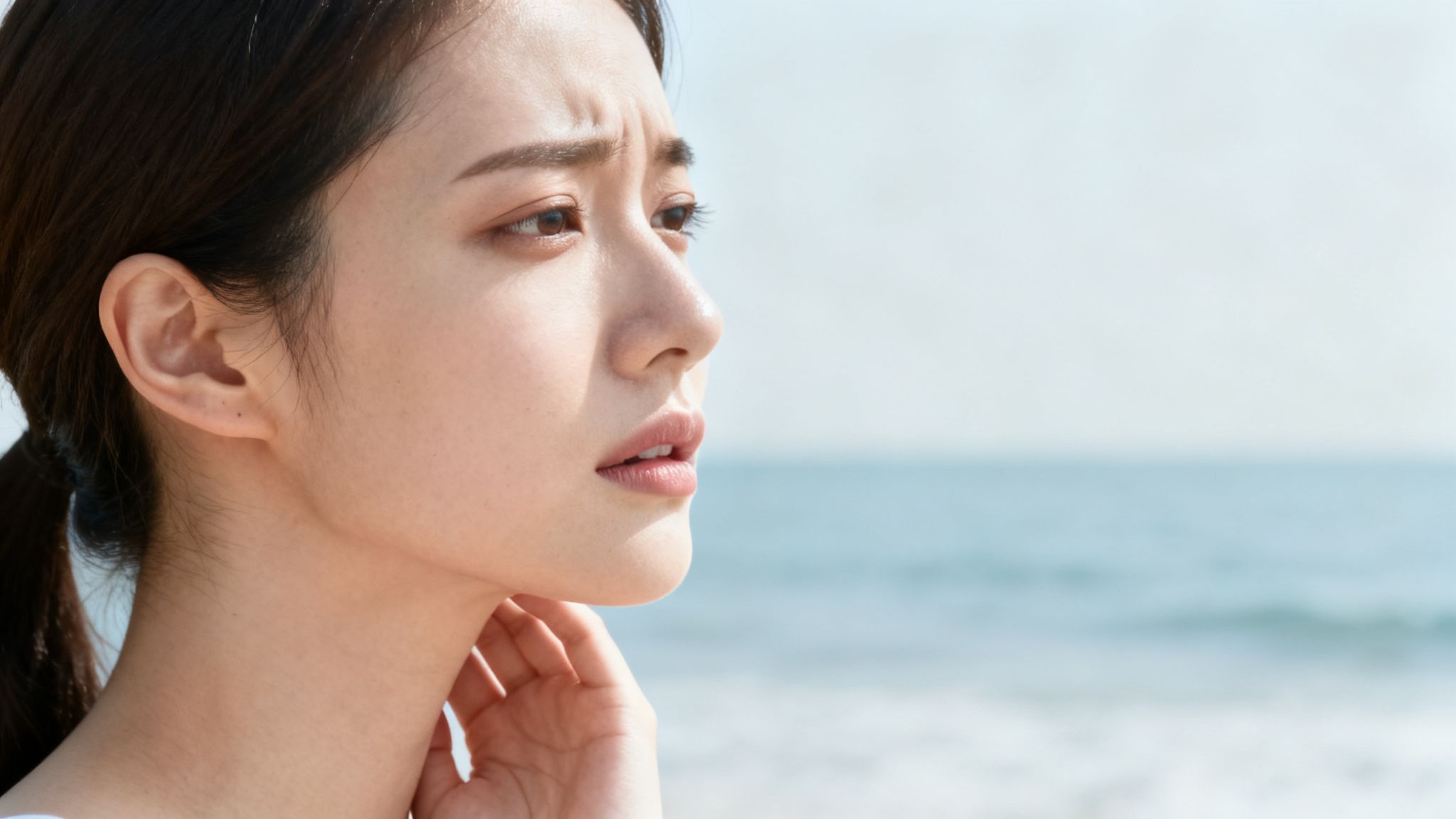 Seitenprofil einer jungen asiatischen Frau am Strand, die besorgt oder nachdenklich aussieht, mit dem Meer im Hintergrund.