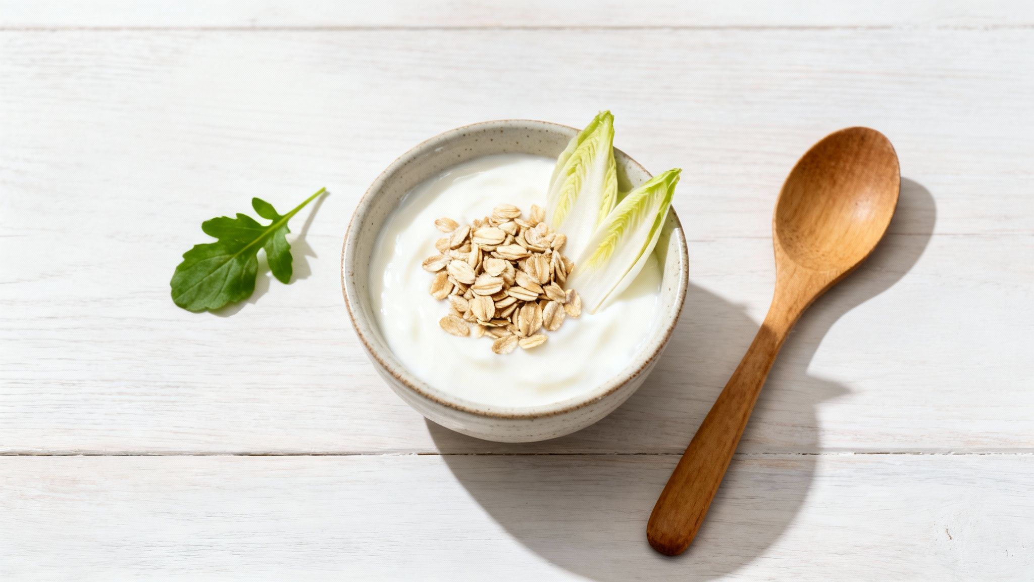 Schale mit Joghurt, Haferflocken und Chicorée auf weißem Holztisch mit Löffel und Blatt.