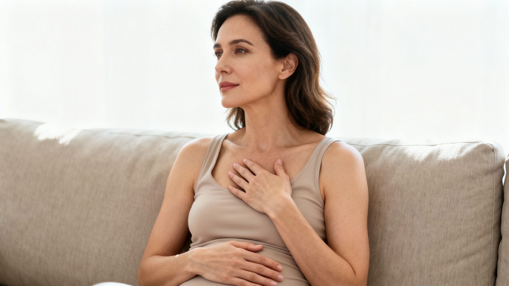 Eine nachdenkliche Frau sitzt auf einem Sofa und hält eine Hand an ihre Brust und die andere an ihren Bauch.
