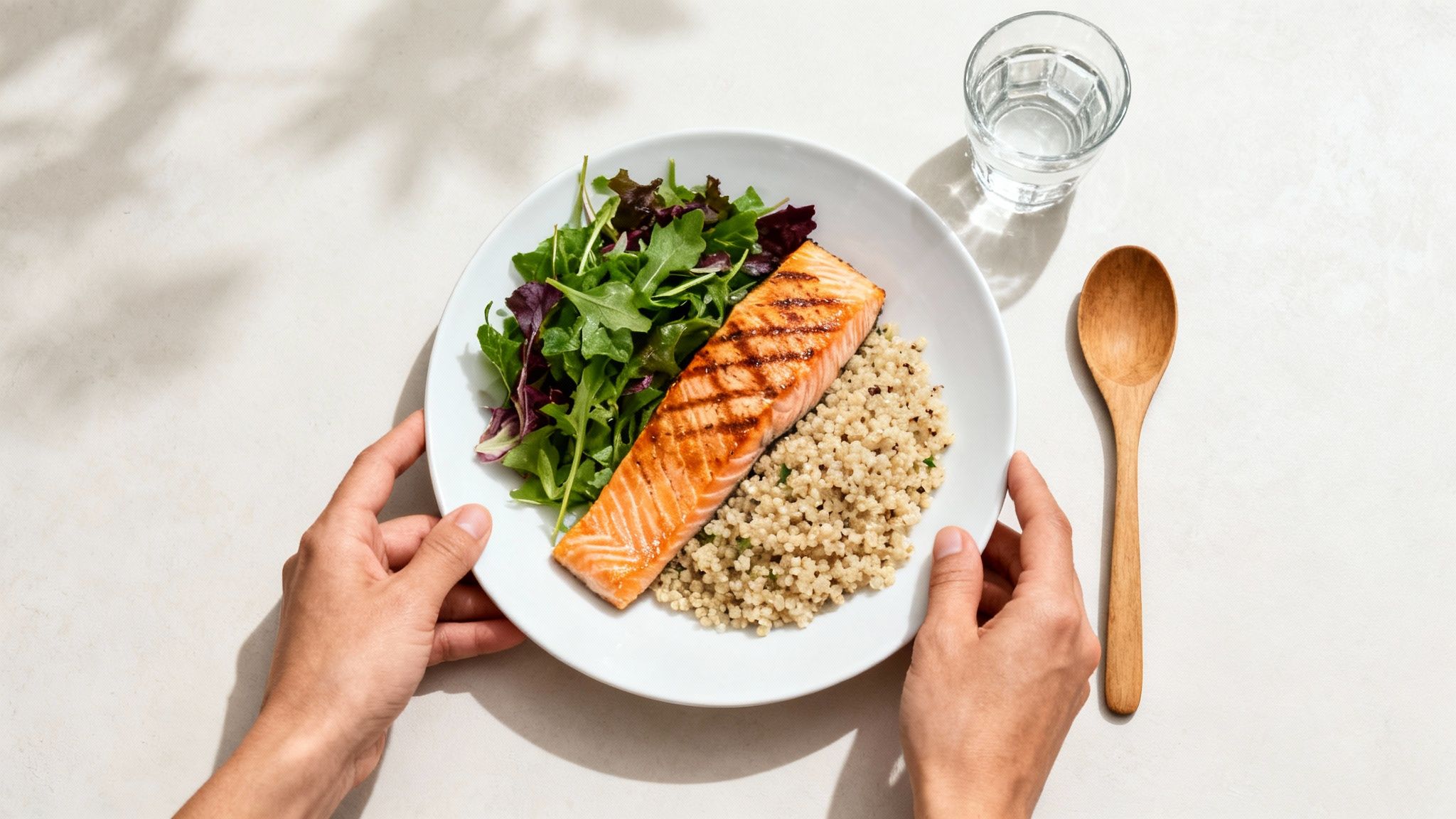 A person is holding a plate with grilled salmon, quinoa and fresh salad, next to a glass of water and a wooden spoon.