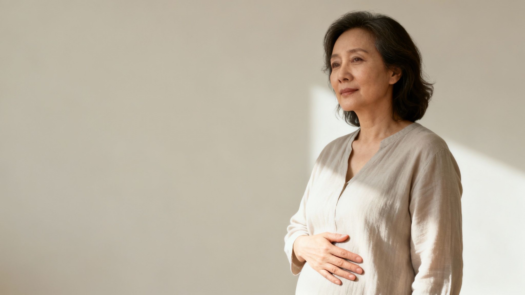 Ältere asiatische Frau mit dunklem Haar, in hellem Hemd, hält ihre Hand auf dem Bauch und blickt nachdenklich in die Ferne.