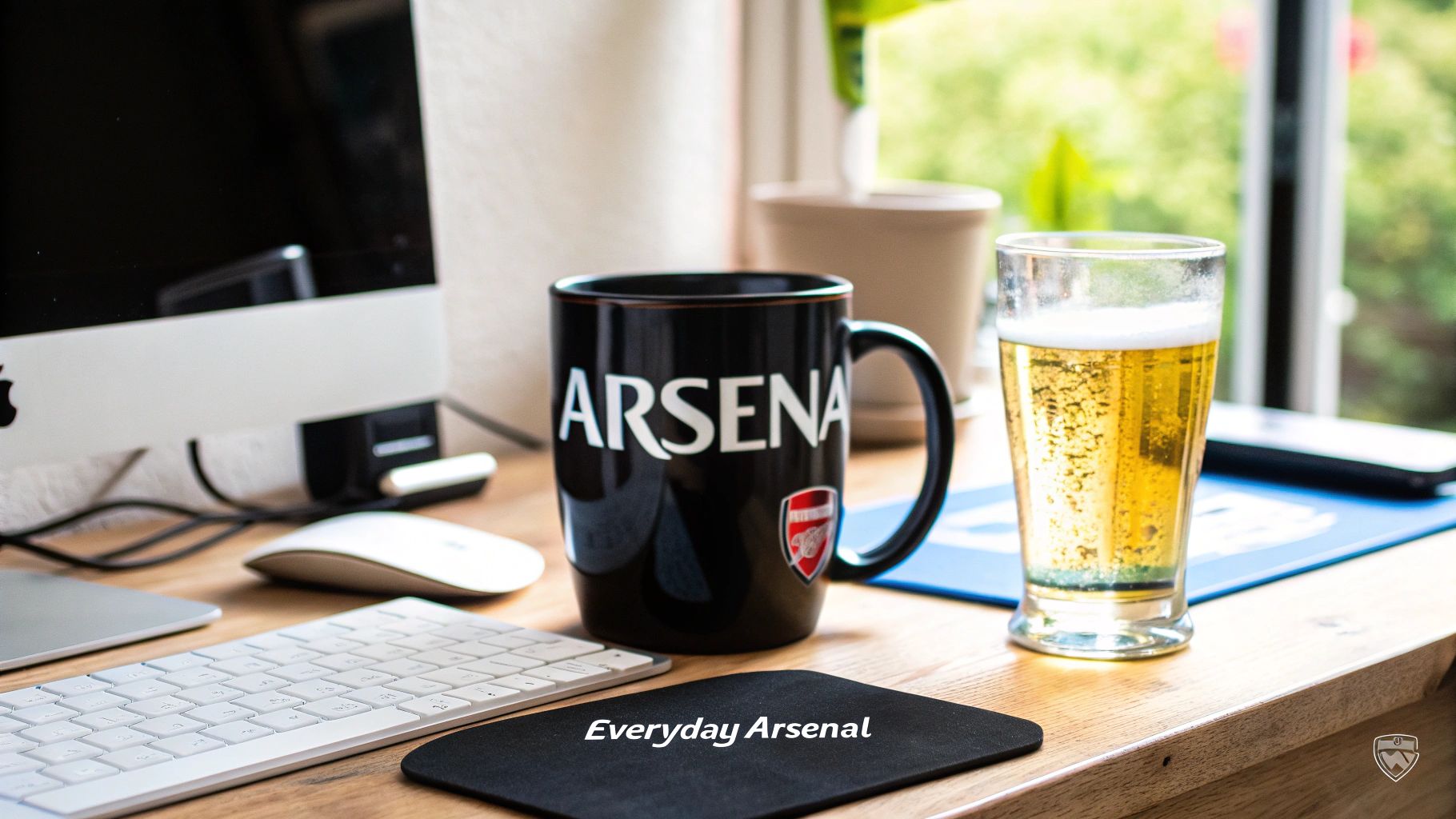 A home office desk with an Apple computer, keyboard, mouse, an Arsenal mug, and a glass of beer.