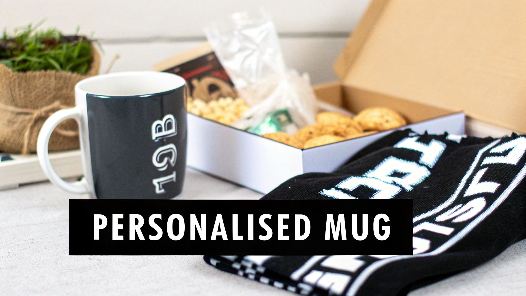 A personalized dark grey and white mug featuring '1GL' text, next to an open gift box with snacks and a scarf.