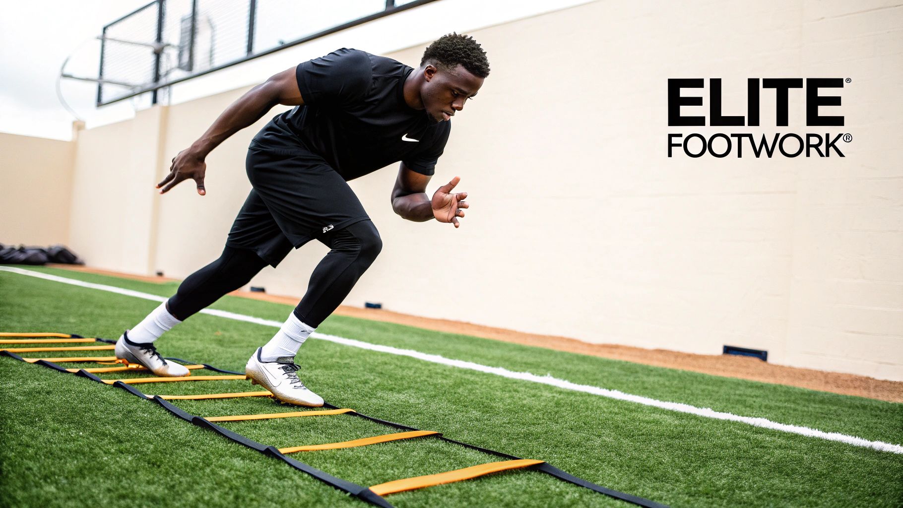 A male athlete in black sports gear performs footwork drills on an agility ladder on a green turf field.