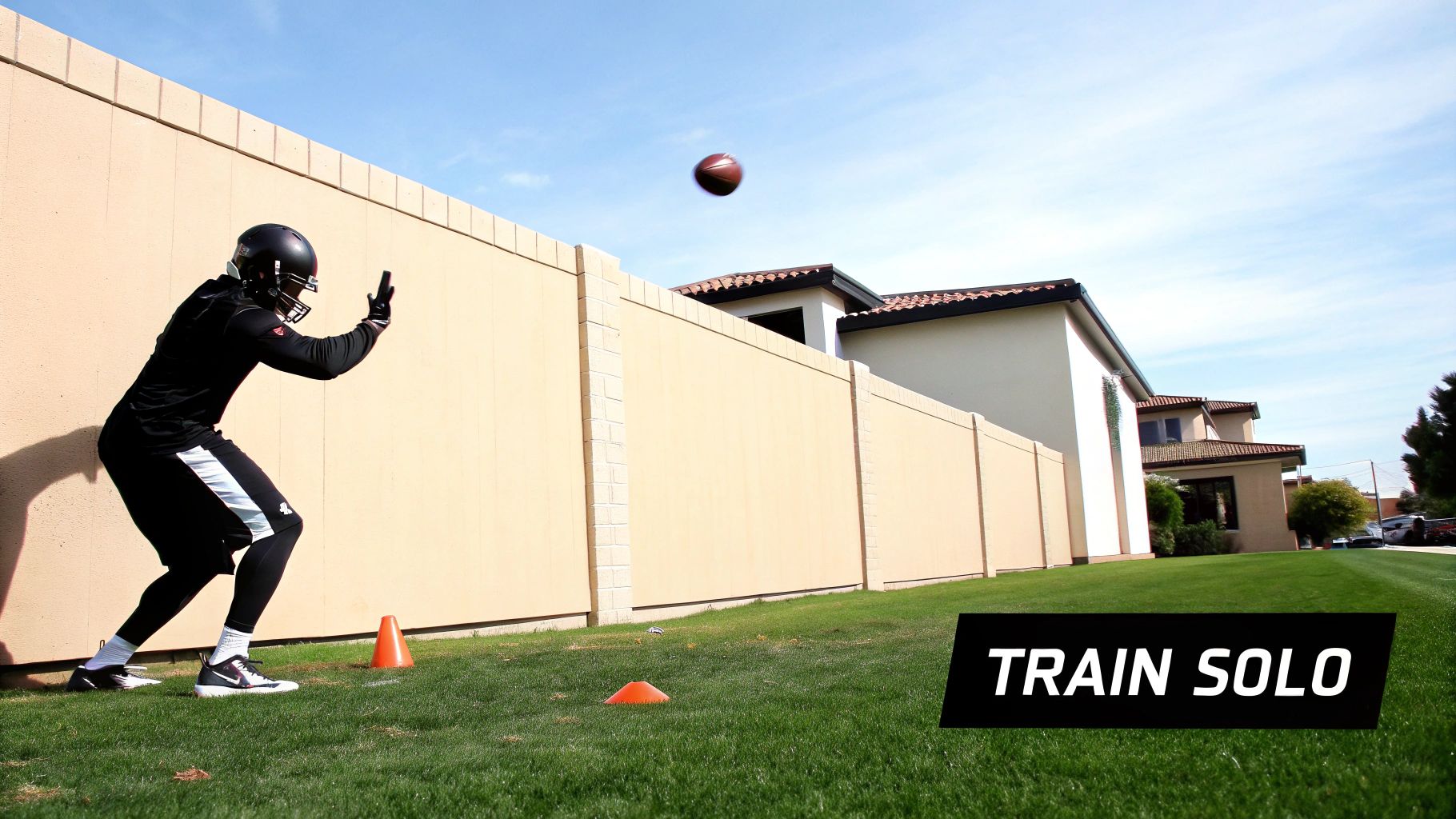 A football player trains solo, catching a football against a wall on a sunny day.