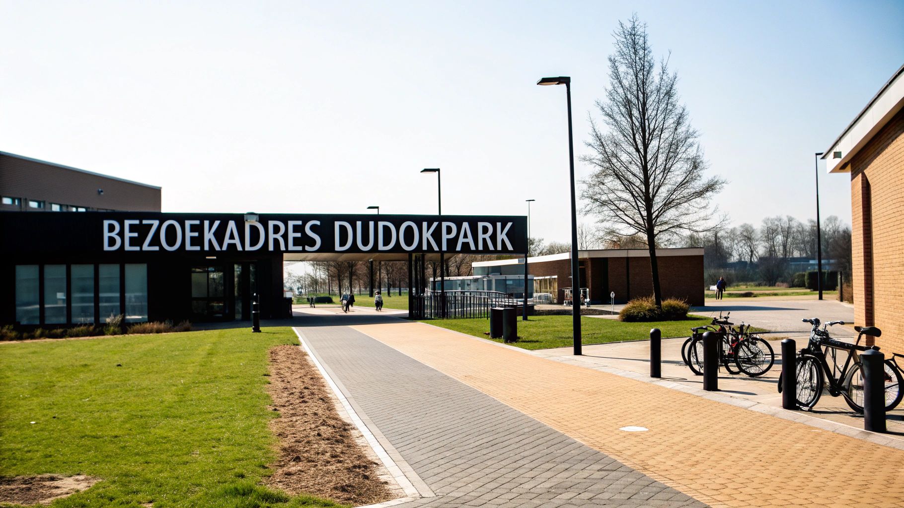 De entree van Dudokpark in Hilversum met een groot zwart bord, een pad en geparkeerde fietsen op een zonnige dag.