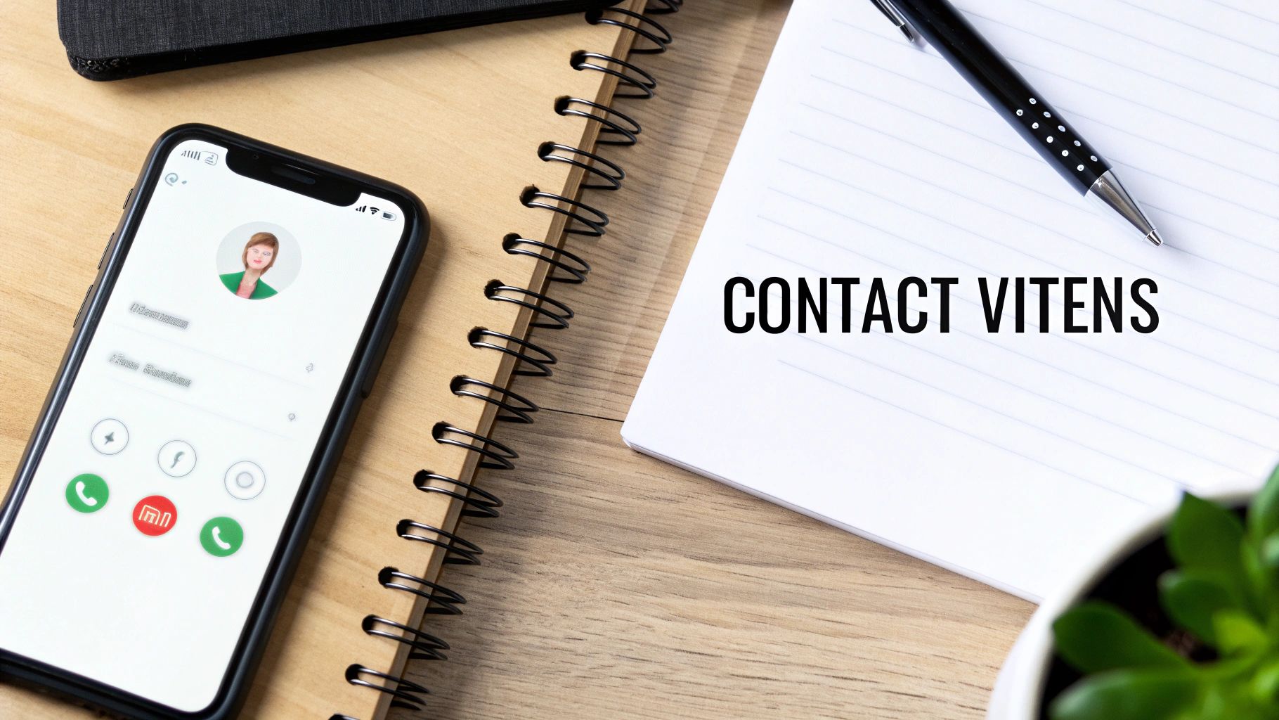 Een smartphone met een contactpersoon en een notitieboekje met de tekst 'CONTACT VITENS' op een houten bureau.