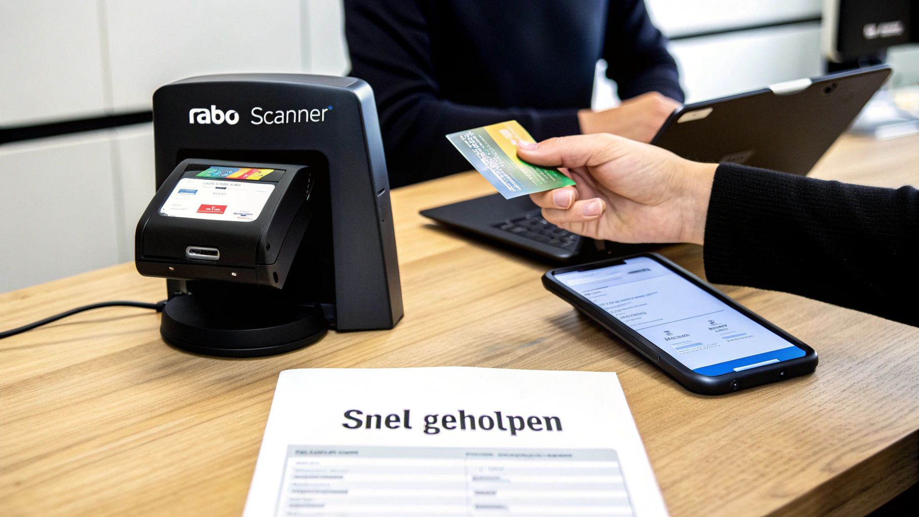 Een hand houdt een bankpas vast bij een Rabo Scanner op een houten tafel, naast een smartphone en document.
