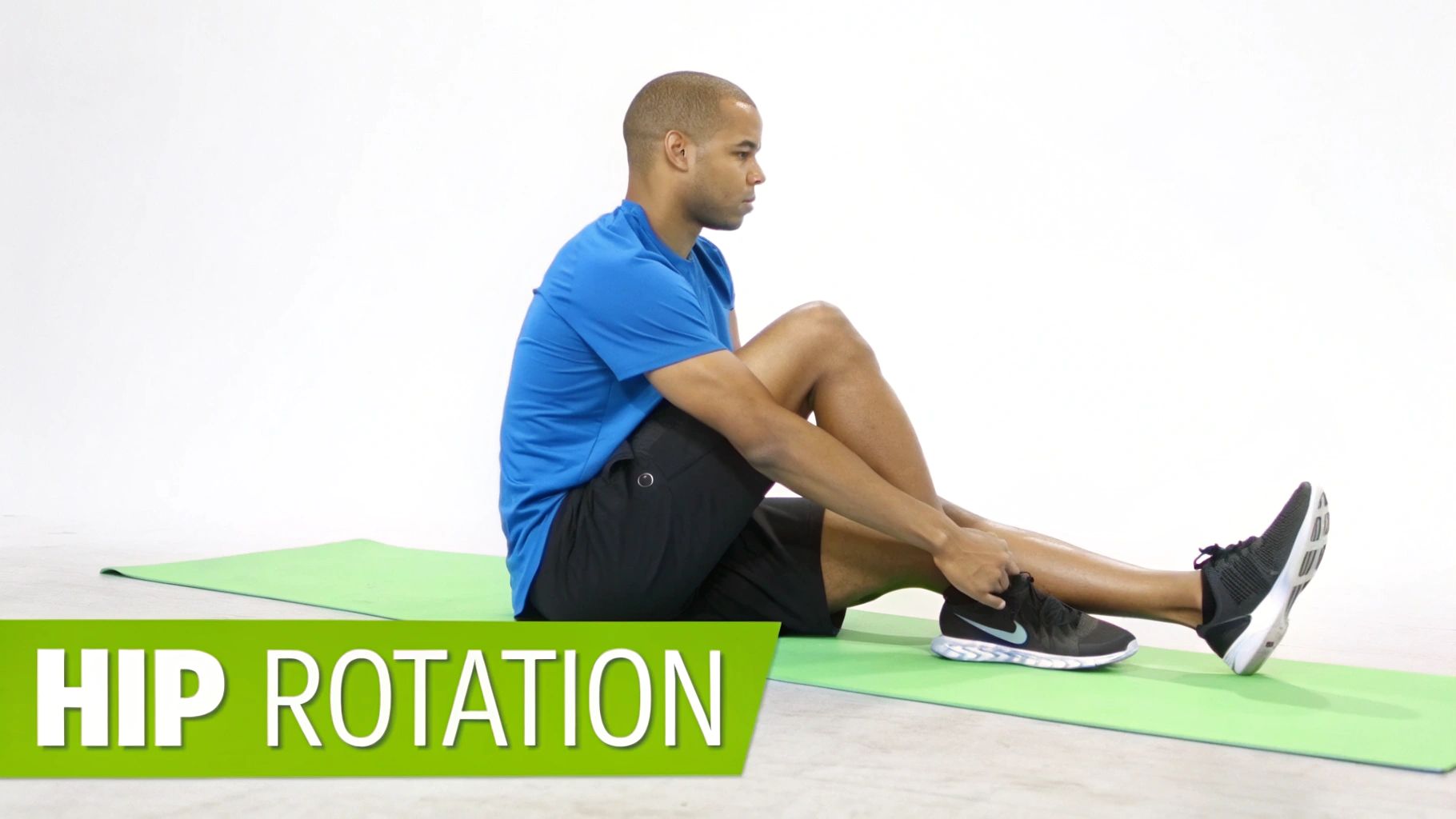 A man sits on a green mat, holding his ankle while performing a hip rotation exercise.