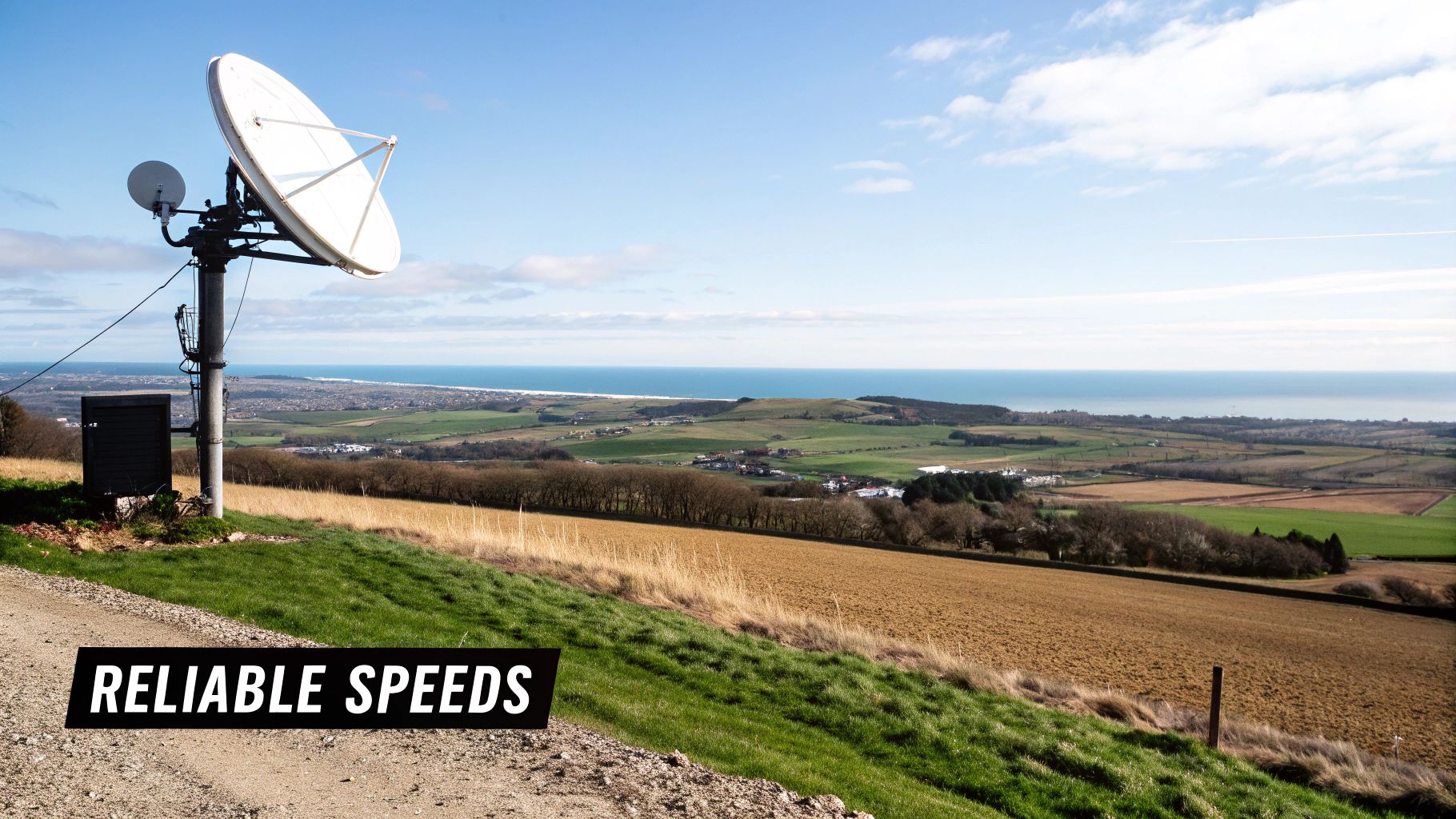 A large satellite dish on a pole on a grassy hill, overlooking a coastal town, ocean, and blue sky.