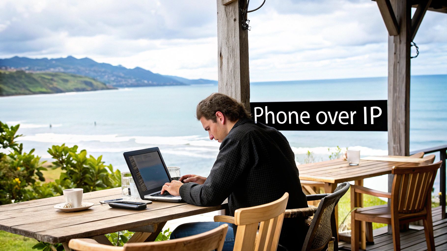 A man working on a laptop at a wooden table outdoors with an ocean and mountain view.
