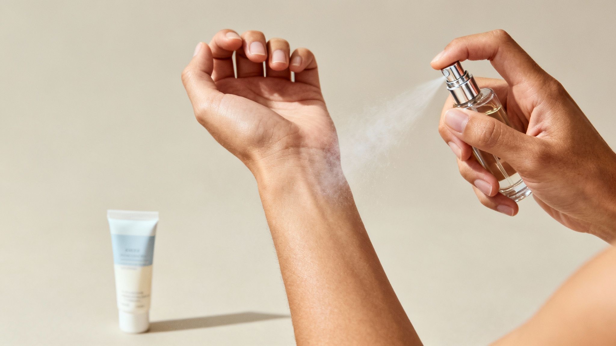 A person sprays a fragrance onto their wrist, creating a visible mist, with a cream tube in the background.