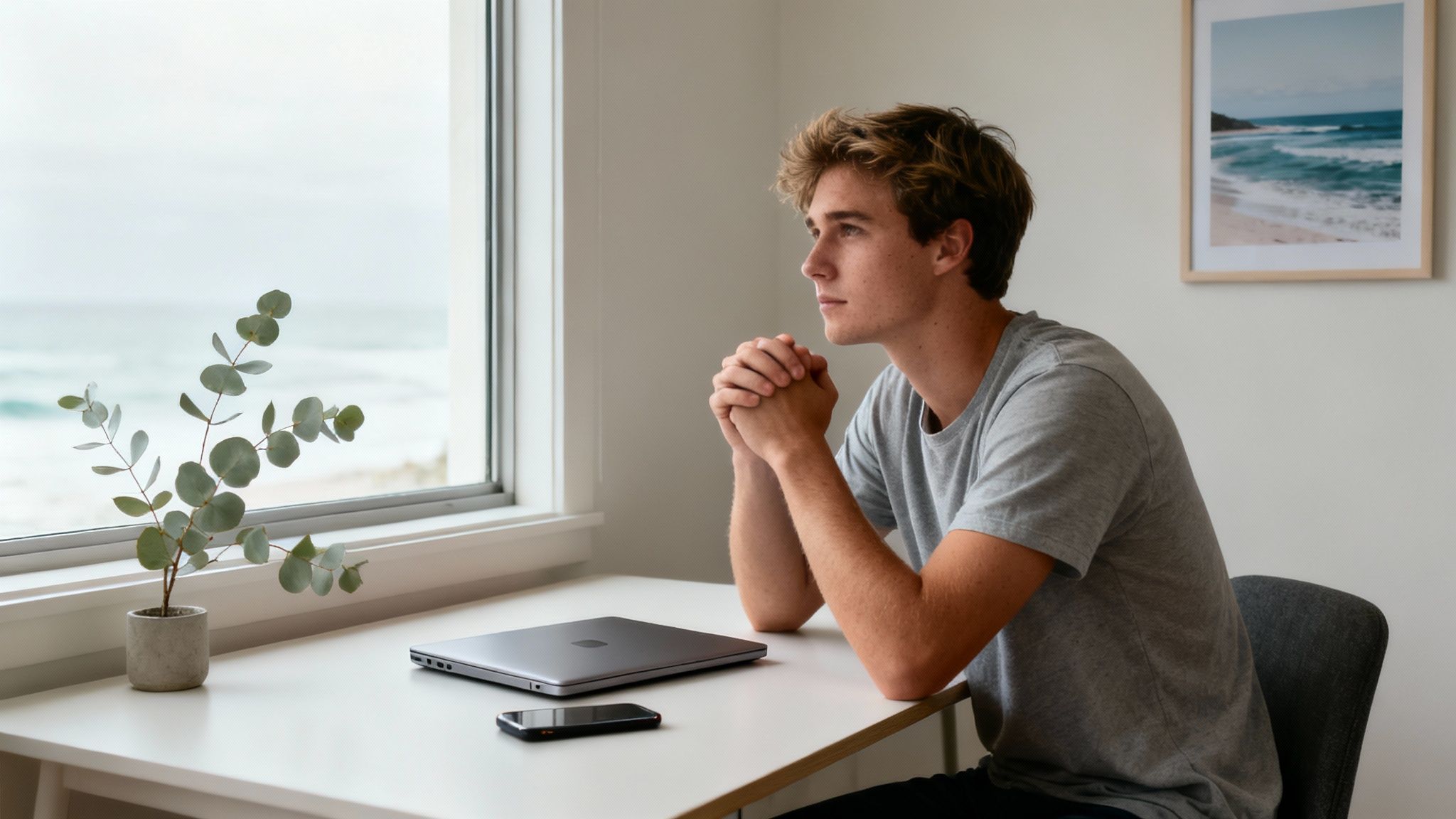 A young man sits at a desk with a laptop, looking thoughtfully out a window at the ocean.