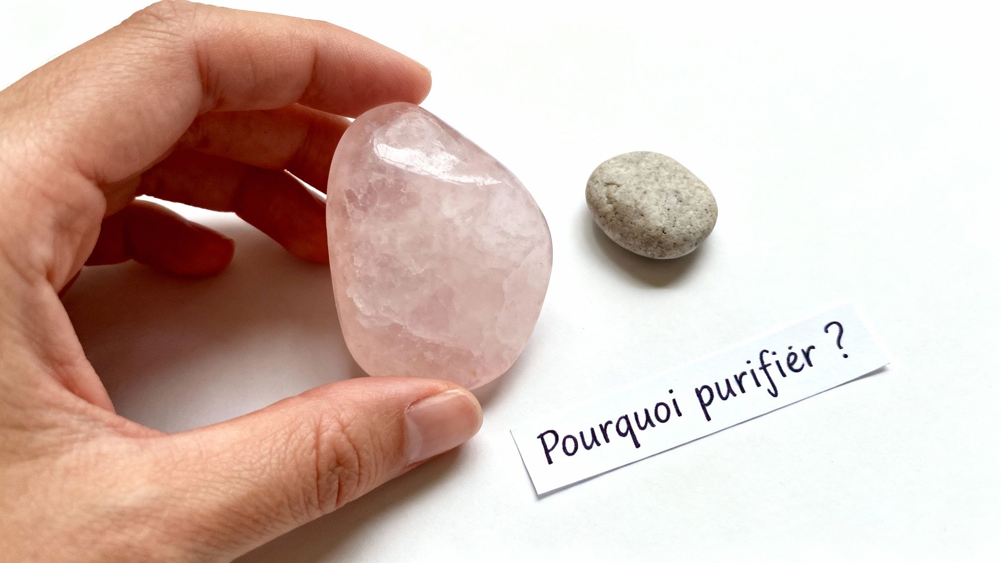 Main tenant une pierre de quartz rose, avec un galet et la question 'Pourquoi purifier ?' sur fond blanc.