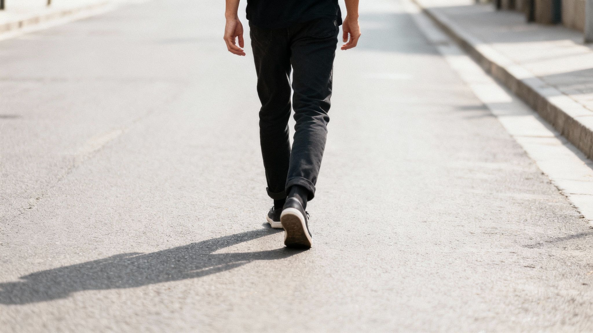 Une personne marche sur une route d'asphalte, vue de dos, avec son ombre projetée.