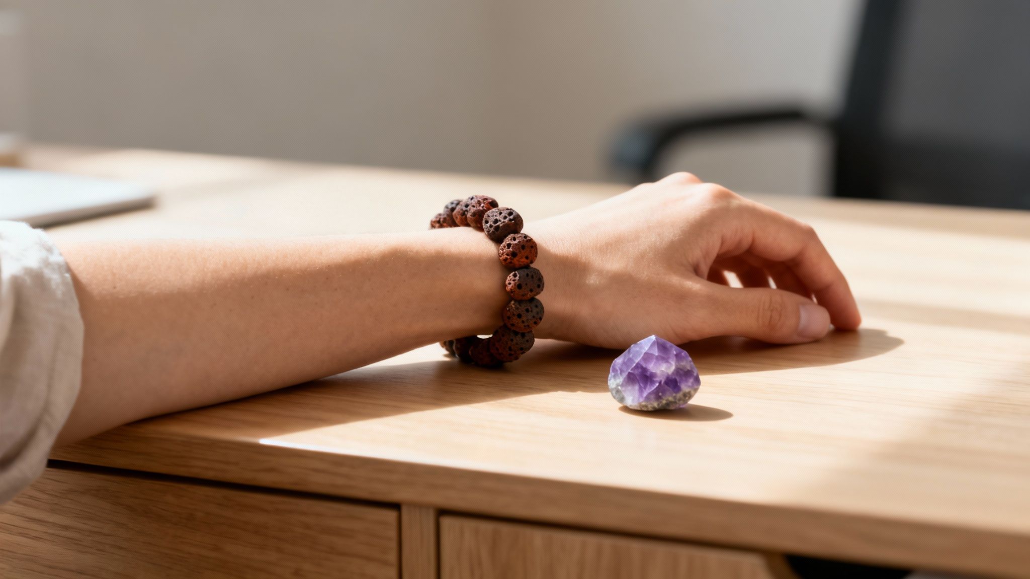 Bras d'une personne portant un bracelet en perles de lave, avec un cristal d'améthyste sur un bureau en bois ensoleillé.