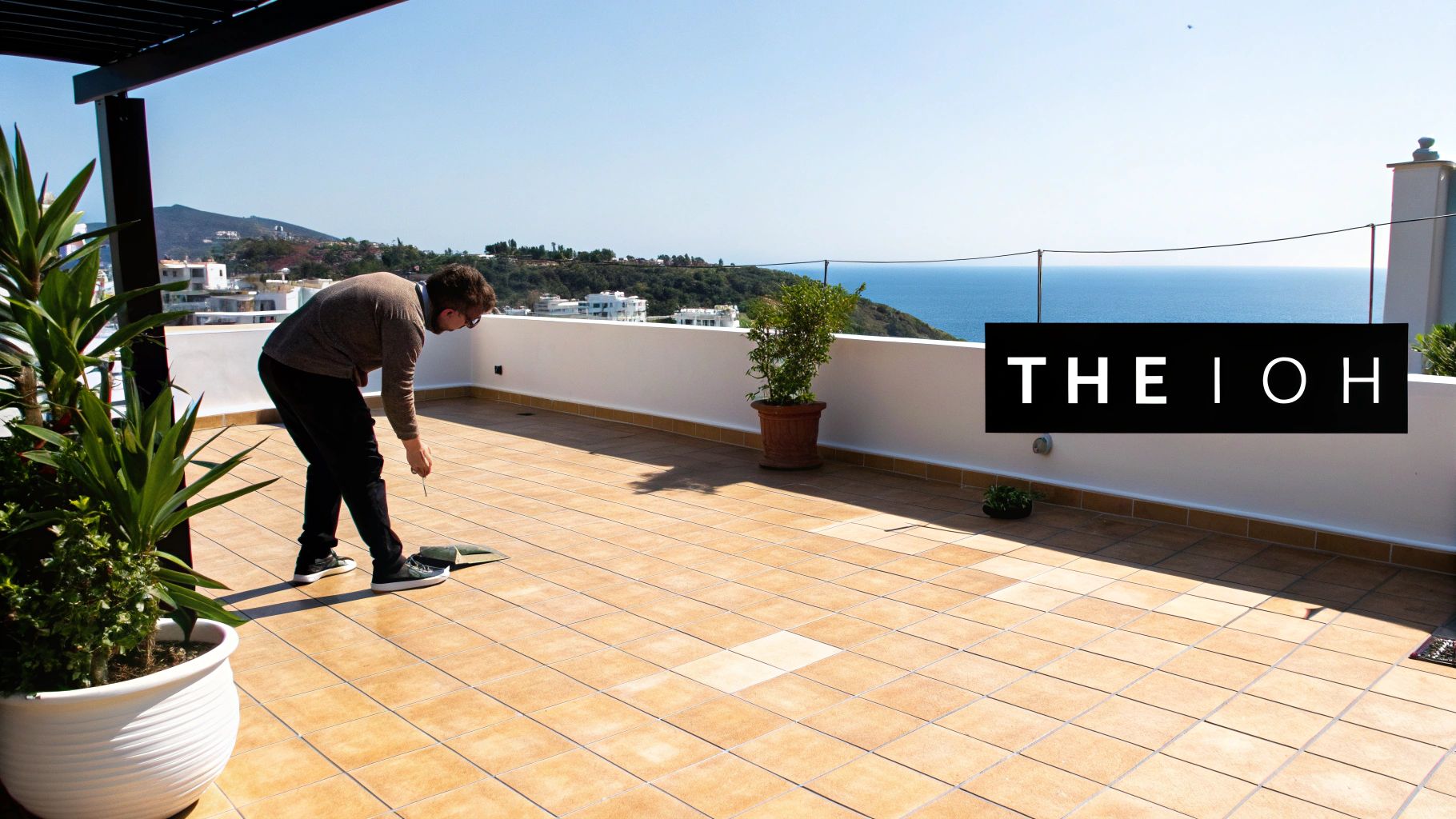 Ένας άνδρας σκύβει σε μια μεγάλη εξωτερική βεράντα με πλακάκια, με εκπληκτική θέα στη θάλασσα και τα βουνά.