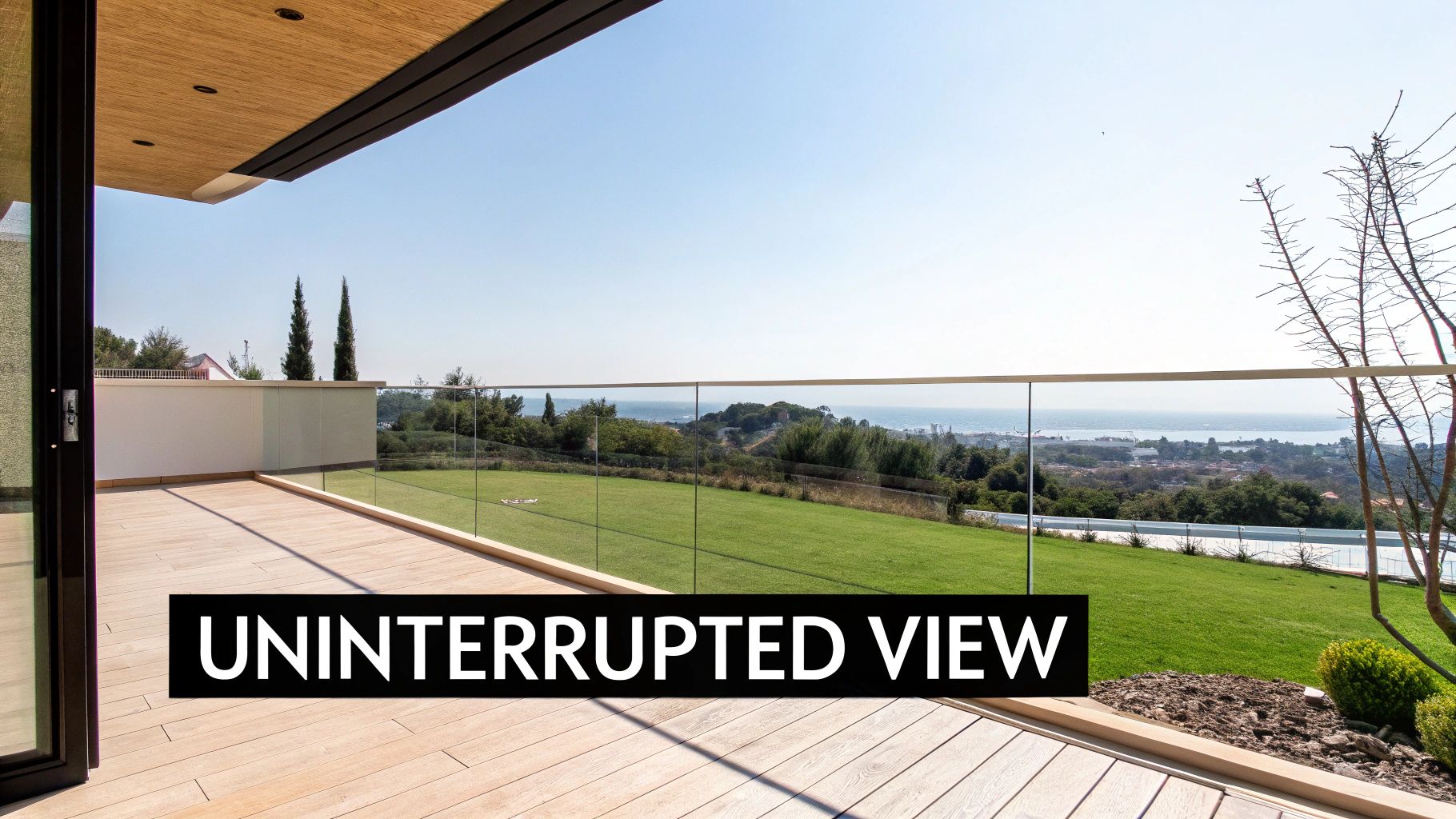 A modern outdoor wooden deck with a glass balustrade offering an expansive, uninterrupted view of a green landscape and the sea.