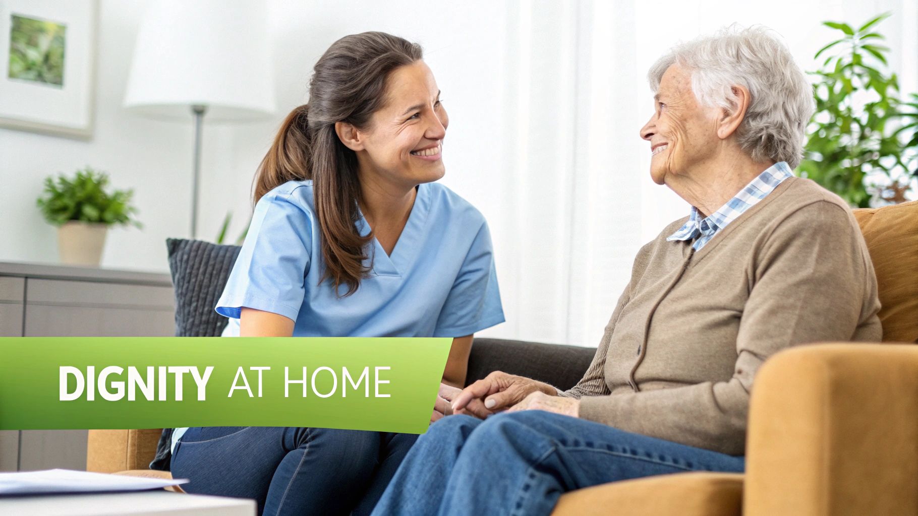 A smiling female caregiver in blue scrubs converses with a happy elderly woman in a home setting.