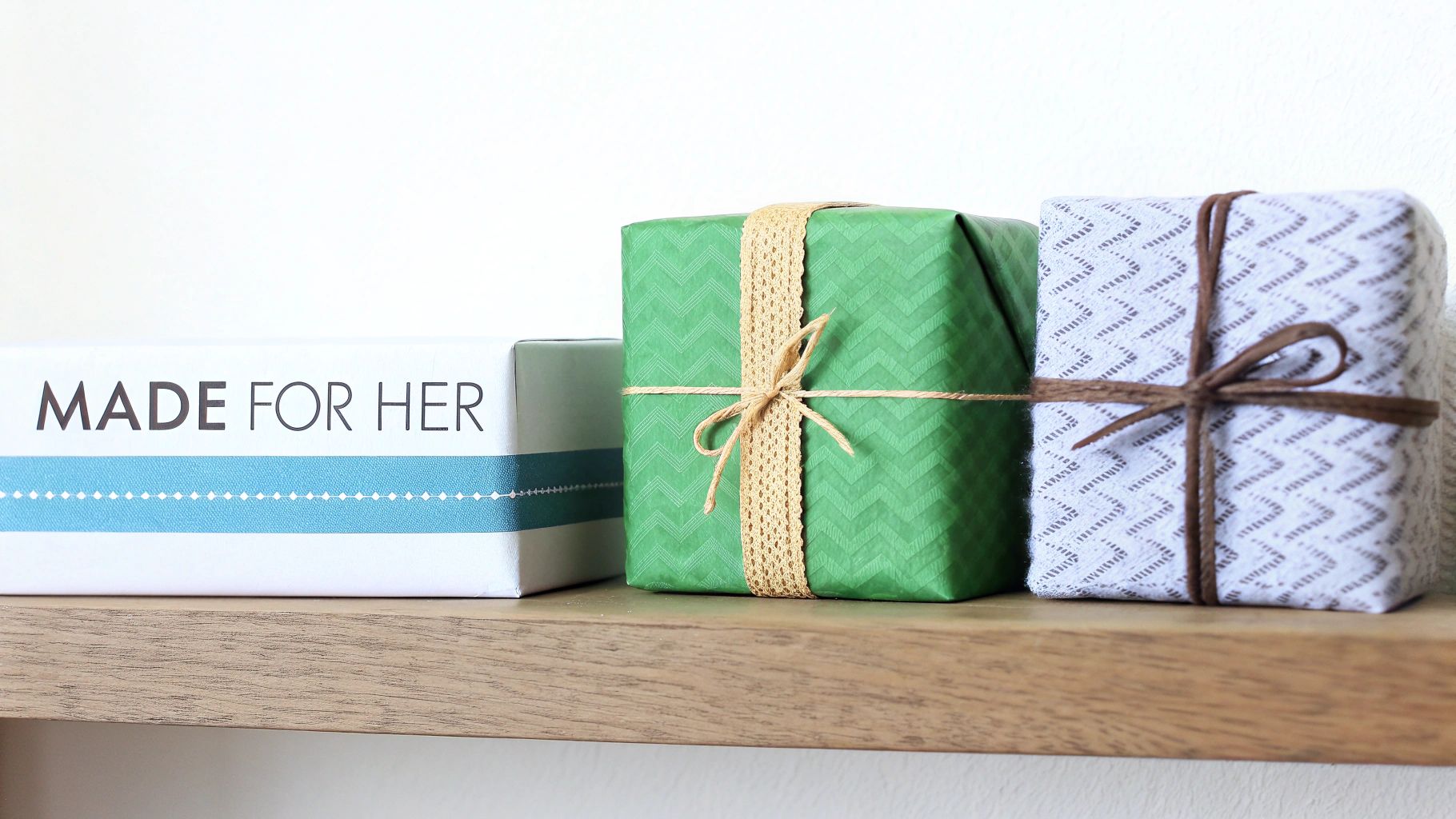 Three beautifully wrapped gifts, including a "MADE FOR HER" box, on a wooden shelf.