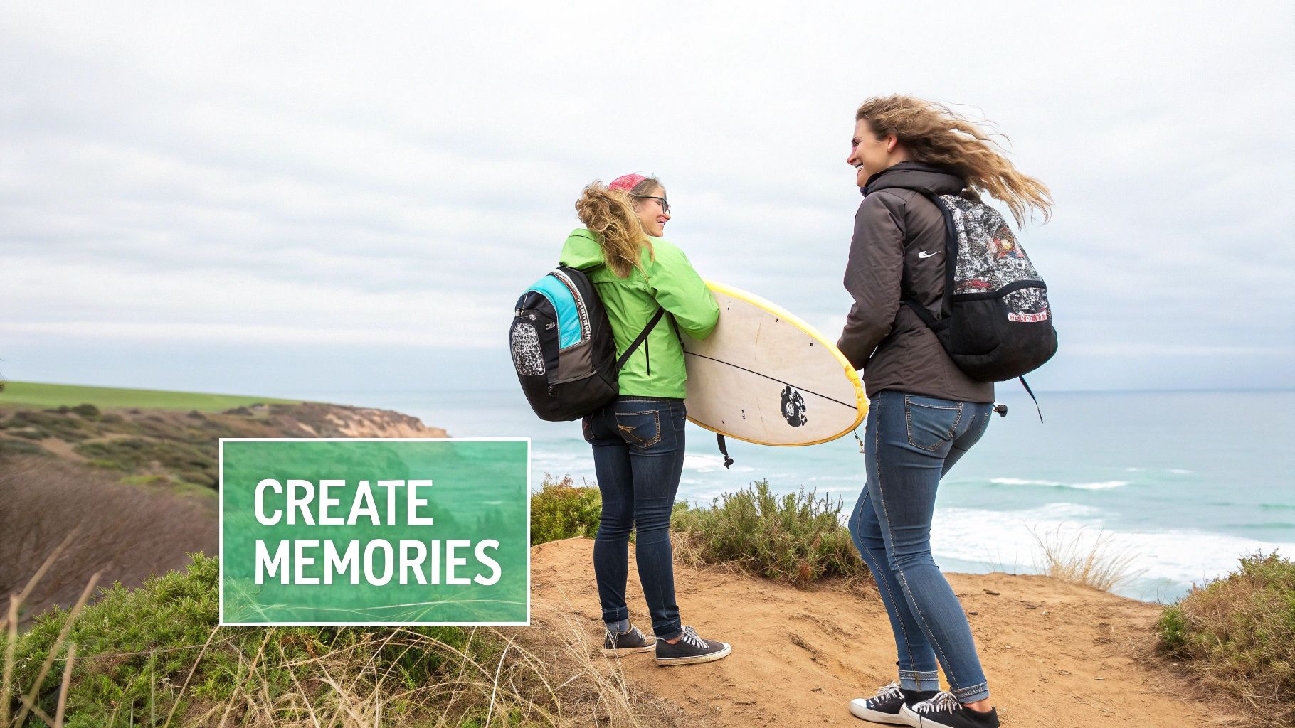 Two happy women with backpacks and a surfboard on a scenic cliff overlooking the ocean.