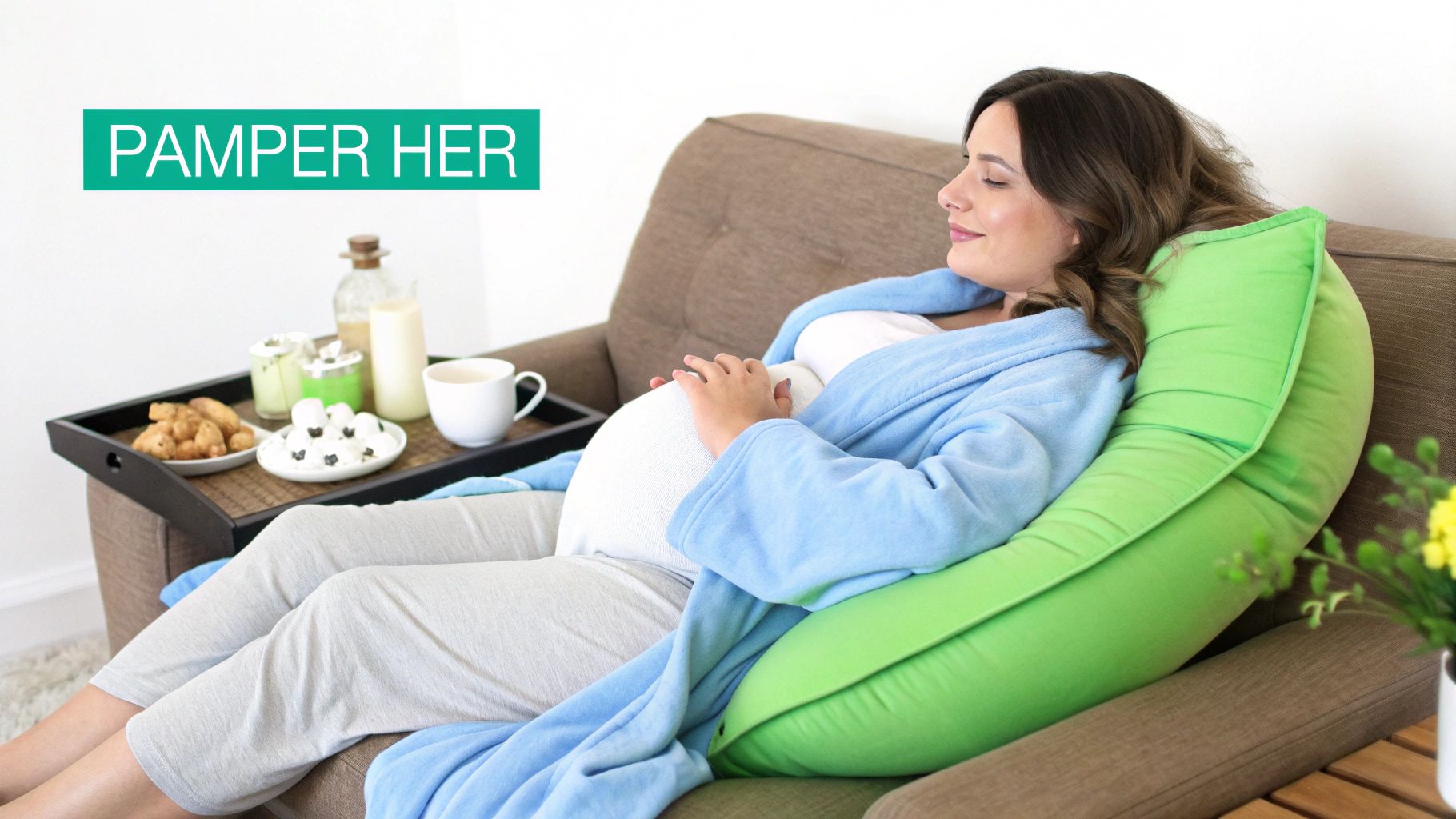A pregnant woman in a blue robe relaxes on a sofa with a green body pillow and snacks.