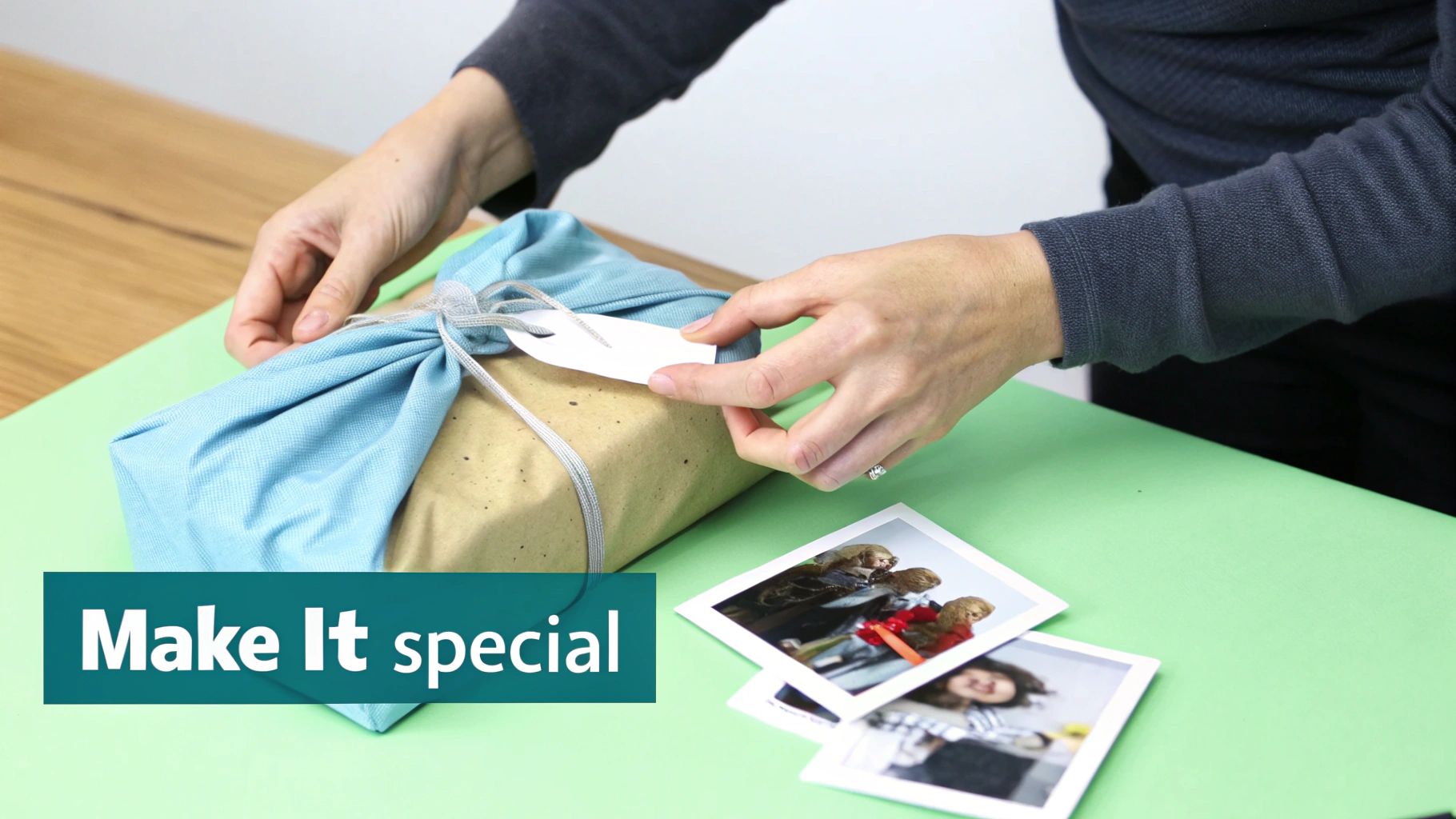 A person's hands attach a white gift tag to a present wrapped in blue fabric, with photos on a green surface.