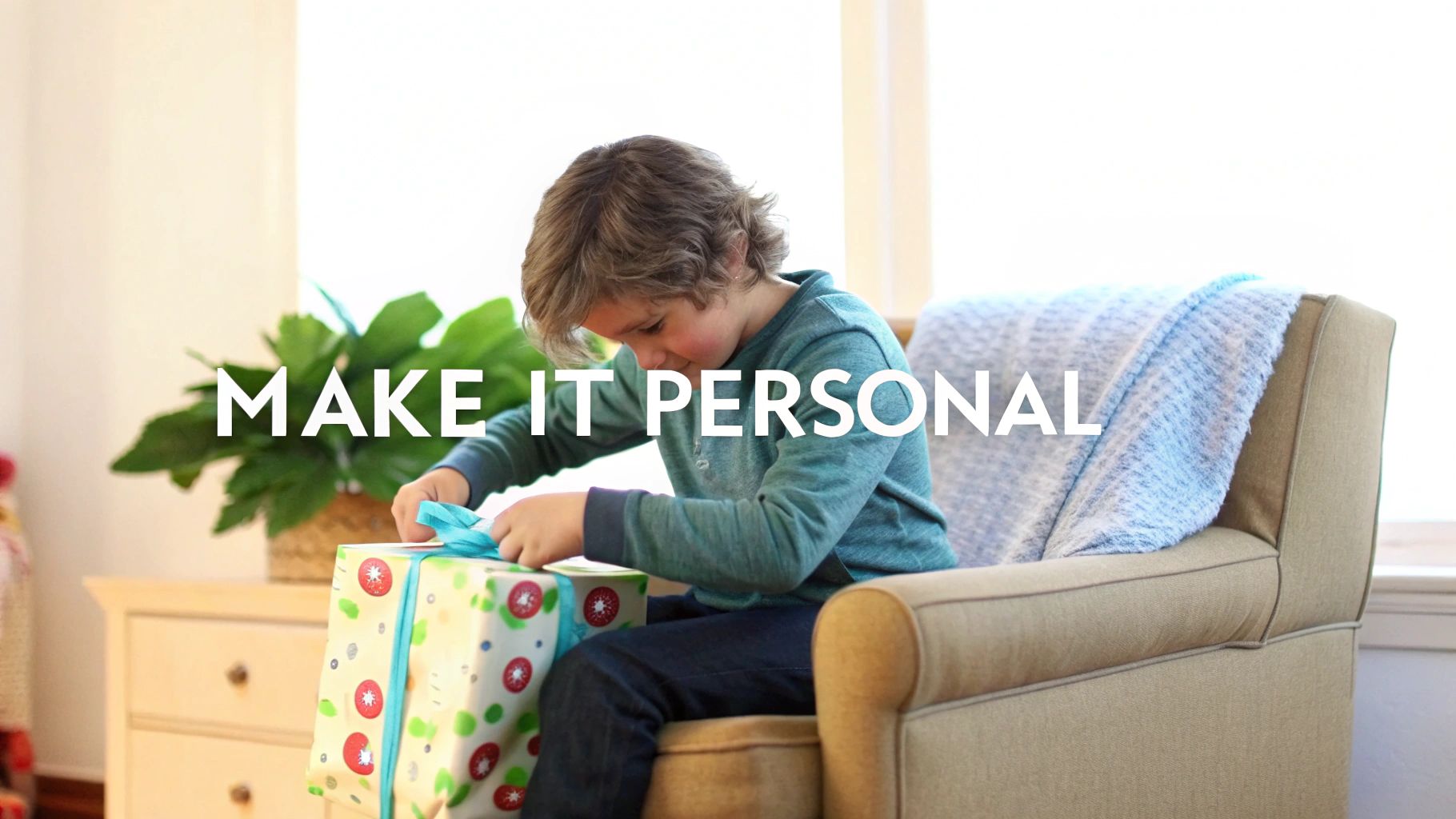 A young boy with curly hair unwrapping a Christmas present with blue ribbon, sitting in an armchair.