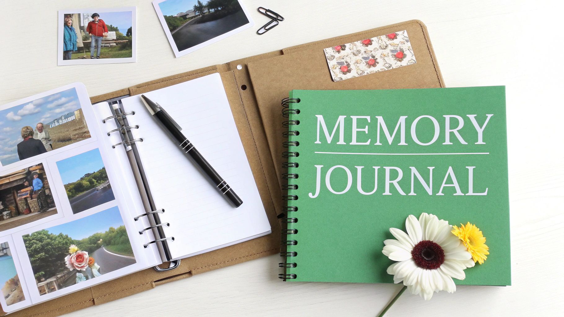 A flat lay of a memory journal, an open photo album with pictures, a pen, and flowers on a white wooden table.