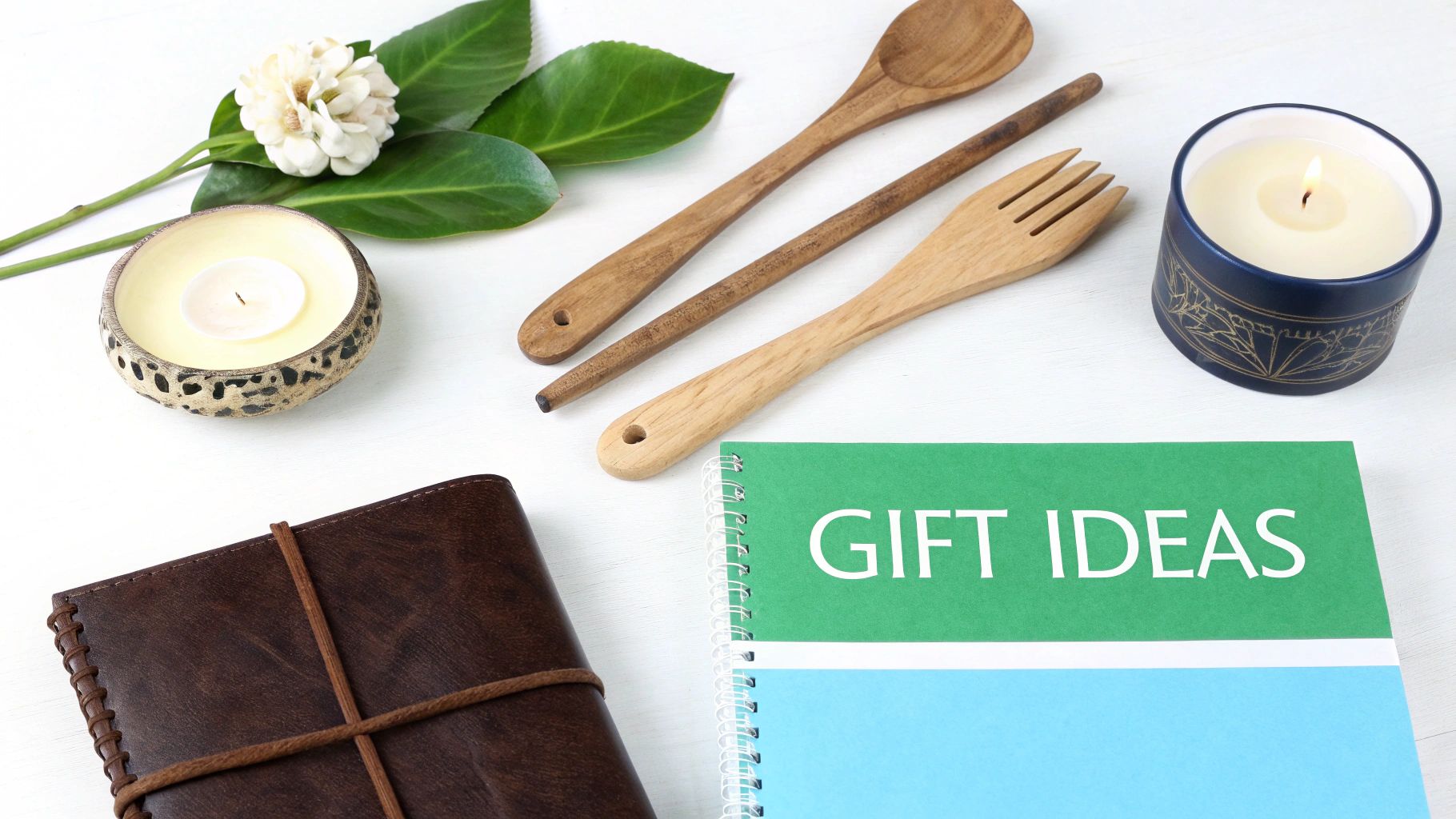 Flat lay of a 'Gift Ideas' notebook, lit candles, wooden utensils, a flower, and a leather journal.