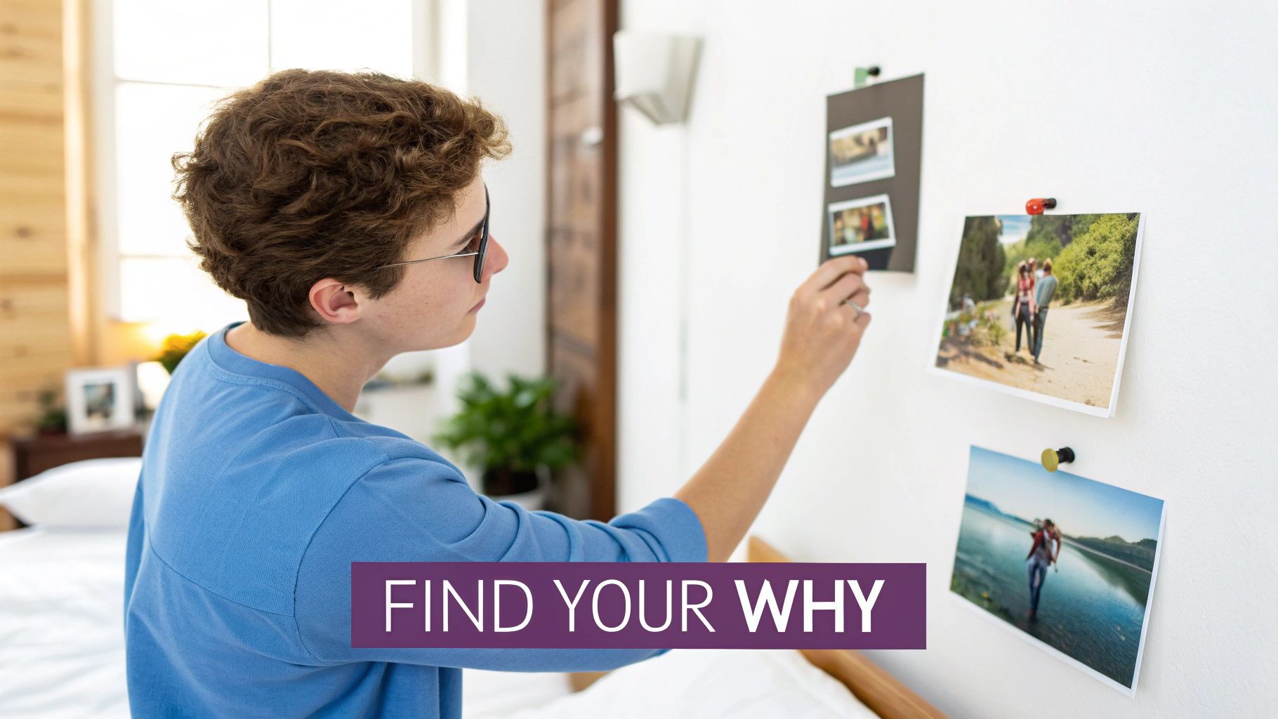 A young person with curly hair and glasses looks at photos pinned to a white wall, creating a vision board.