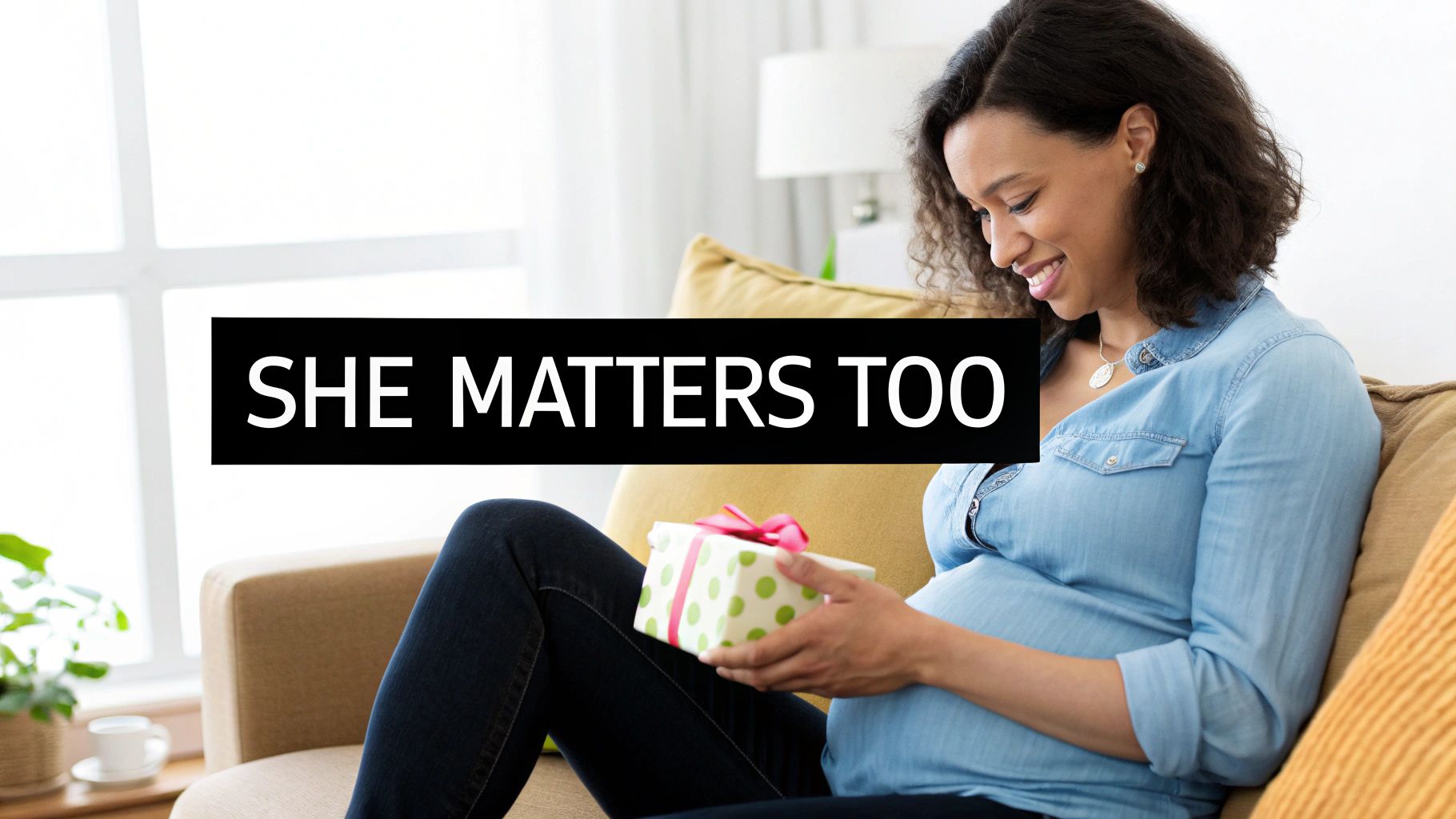 A smiling pregnant woman sits on a couch, gently holding a polka-dotted gift box.