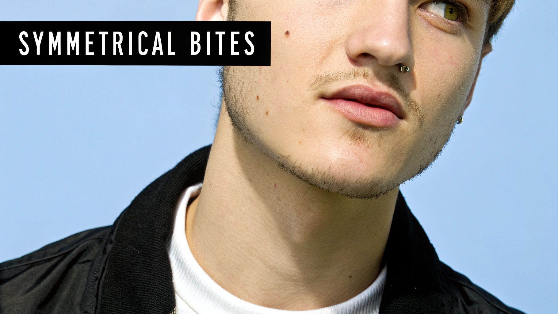 Close-up of a person with a septum piercing and light facial hair, looking upwards against a blue sky.