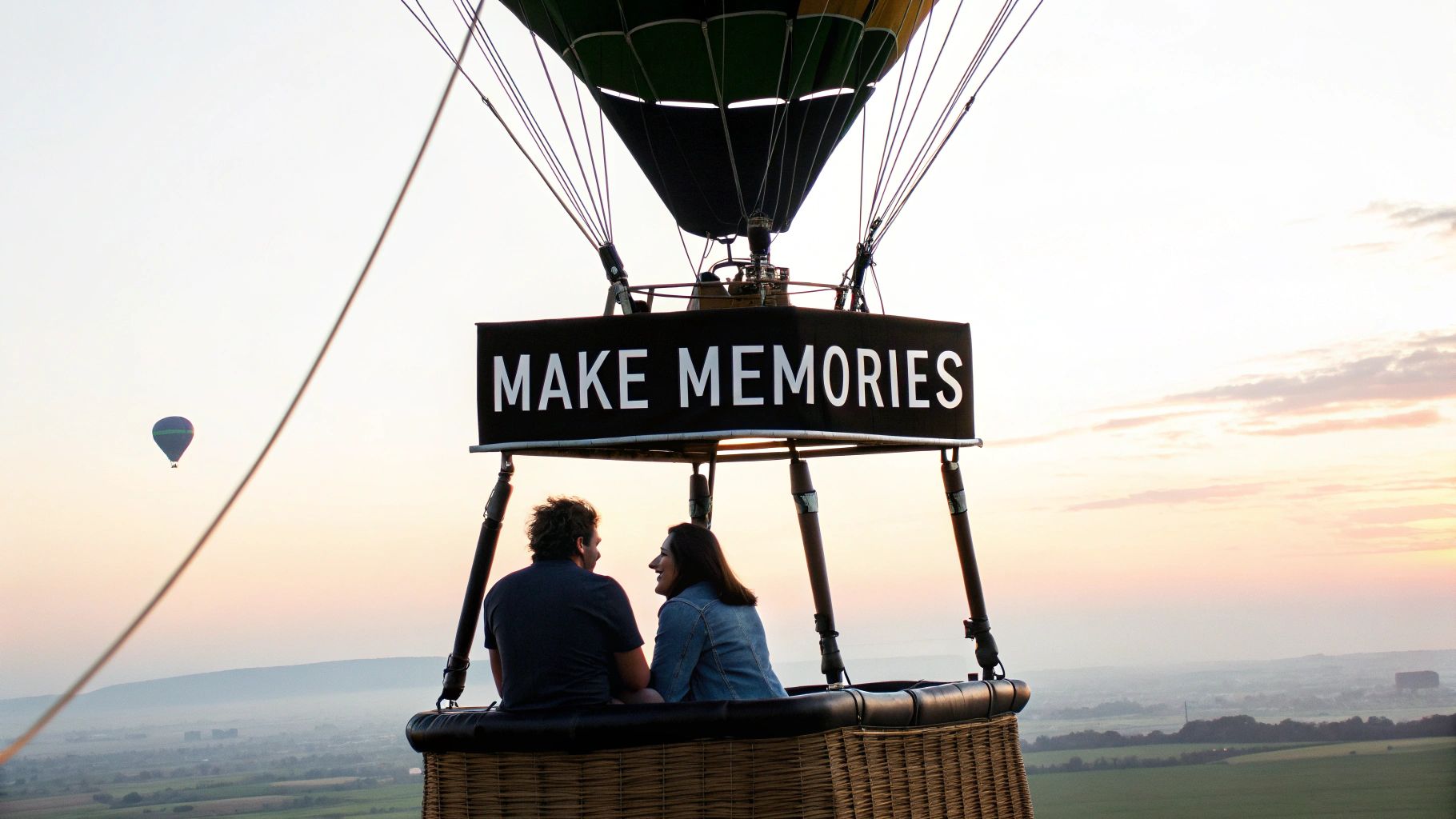 Two people enjoy a hot air balloon ride with a banner saying 'MAKE MEMORIES' over a scenic landscape.