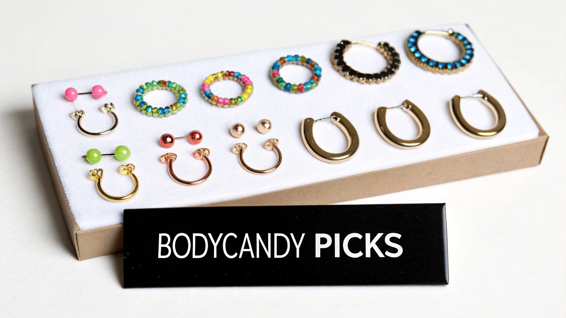 A collection of colorful body jewelry, including barbells, horseshoe rings, and hoop earrings, displayed in a box with 'BODYCANDY PICKS' text.