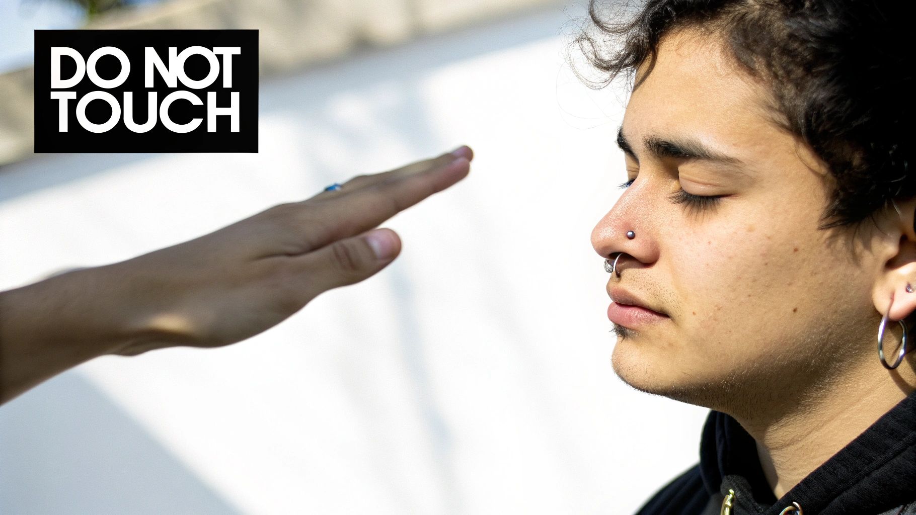 A hand reaches towards a person with nose piercings and closed eyes, next to a 'DO NOT TOUCH' sign.