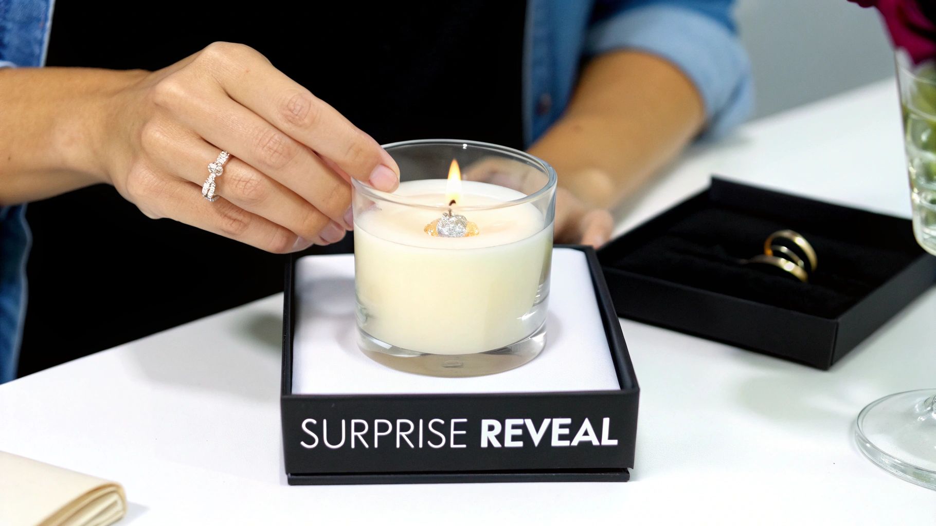 A person's hand holding a lit candle with a diamond ring inside, on a 'SURPRISE REVEAL' box.
