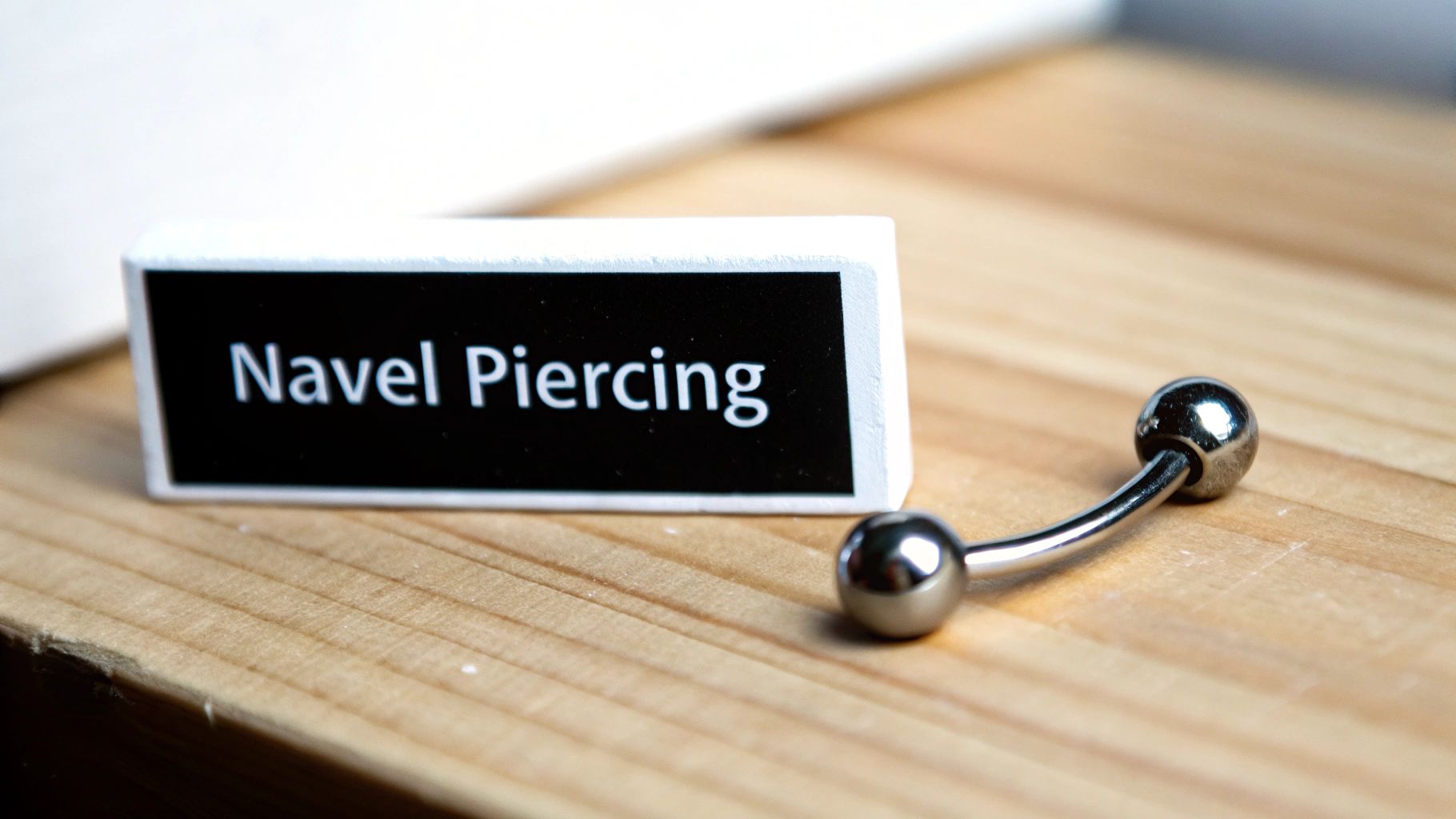 A close-up of a 'Navel Piercing' sign and a silver curved barbell on a wooden table.